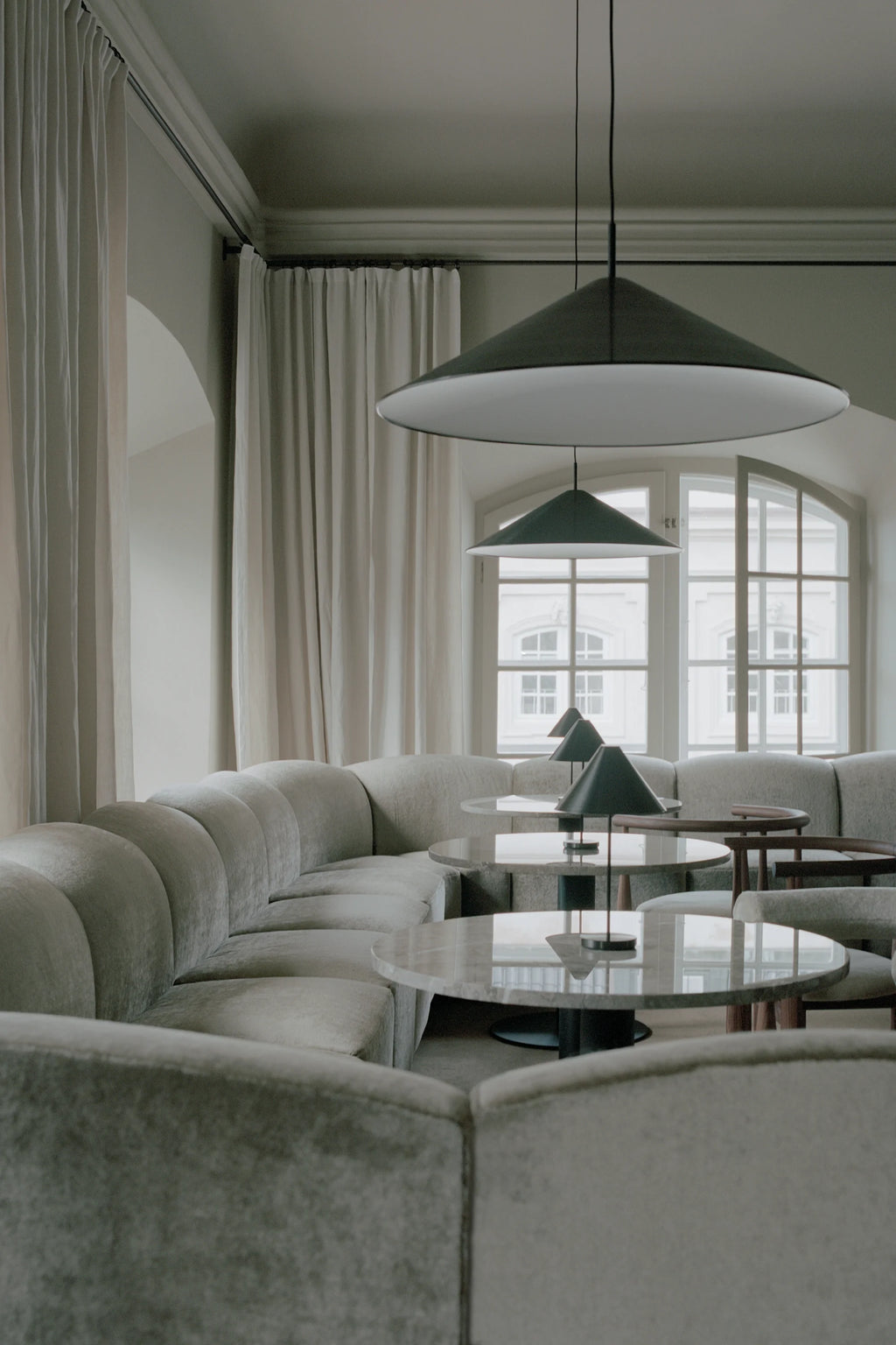 New Works Shore Modular Dining Sofa, a contemporary curved modular sofa upholstered in a soft, textured light grey fabric, featuring a distinctive rounded backrest with vertical channel tufting for a plush, inviting look. This organic modern sectional seating offers flexible arrangements around round marble dining tables, perfect as a stylish banquette for restaurant dining, hotel lounges, or spacious minimalist living rooms.