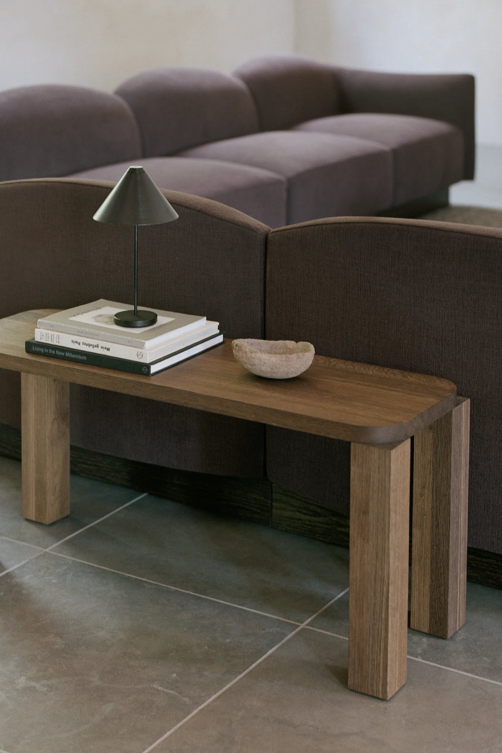 New Works Atlas Bench, a modern minimalist bench crafted from solid dark stained oak wood, featuring a rectangular top with softly rounded edges and thick, sturdy block legs that highlight the natural wood grain. This organic modern oak bench is perfect as a contemporary entryway console, a sleek living room accent, or a functional bedroom furniture piece.
