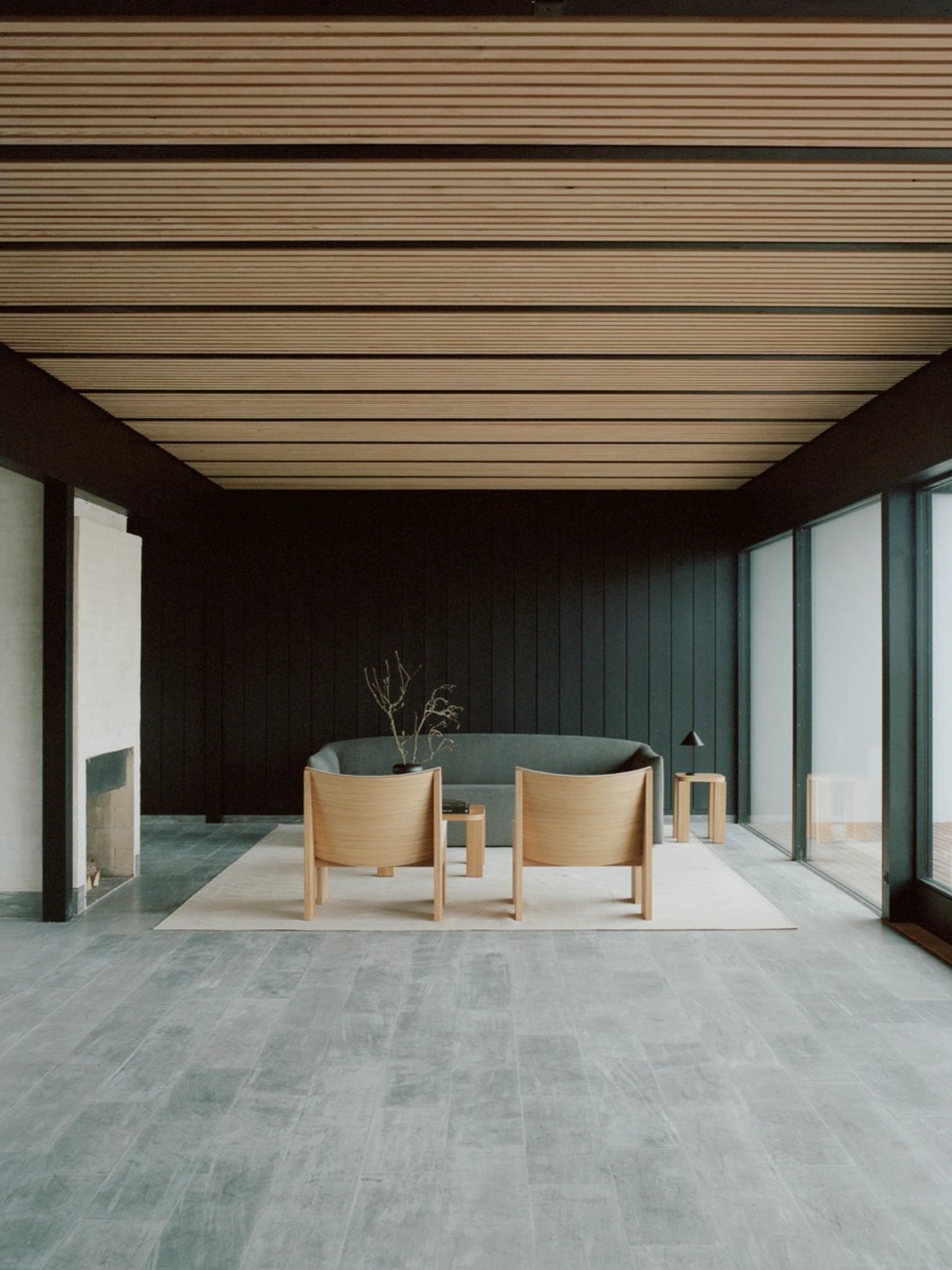 New Works Shore Sofa 3-Seater - Extended Base, in a muted green fabric, anchors a spacious, contemporary living area with dark walls, light wood slatted ceiling, and two elegant wooden lounge chairs facing it.