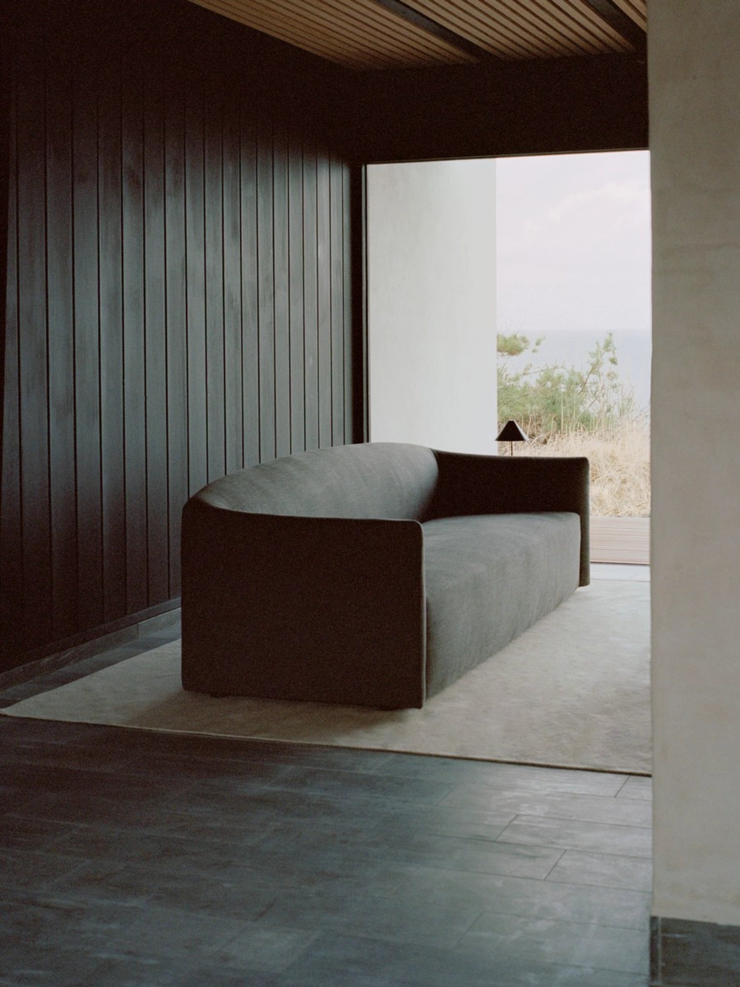 New Works Shore Sofa 3-Seater - Extended Base, in a sophisticated dark grey fabric, with a sleek, curved silhouette, positioned in a modern room with dark wood paneling and a large window overlooking a scenic outdoor view.