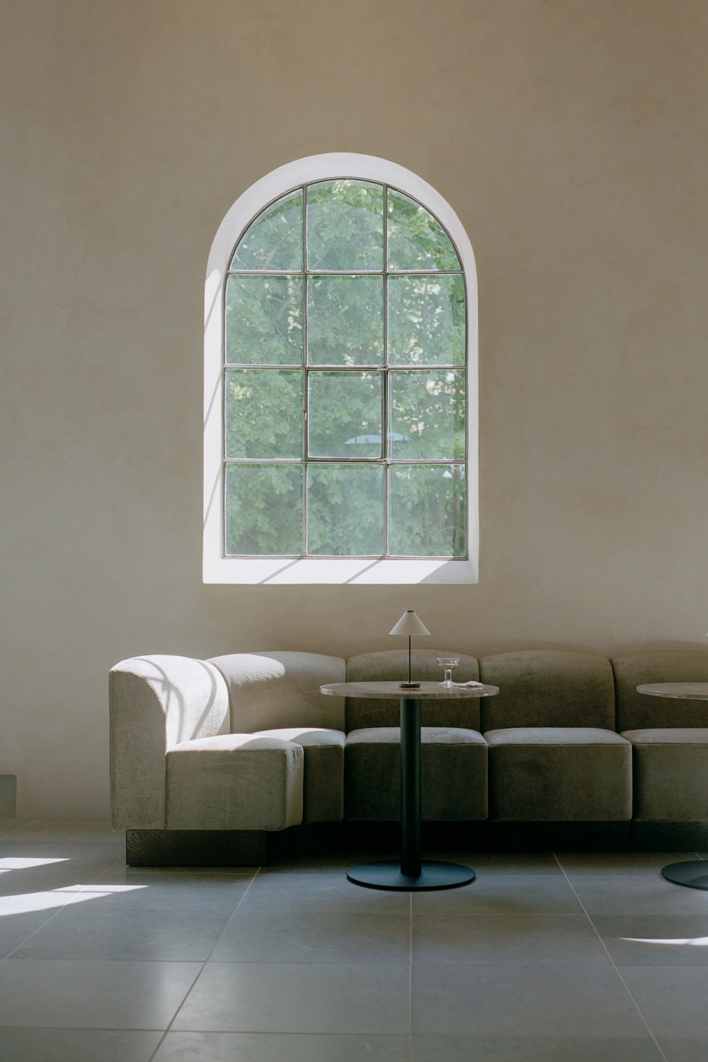 New Works Shore Modular Dining Sofa, a contemporary organic modern sofa featuring soft, textured light beige upholstery with gently curved backrests and a sturdy dark wood block base. This versatile modular seating system includes integrated round light grey marble side tables on matte black metal pedestals.