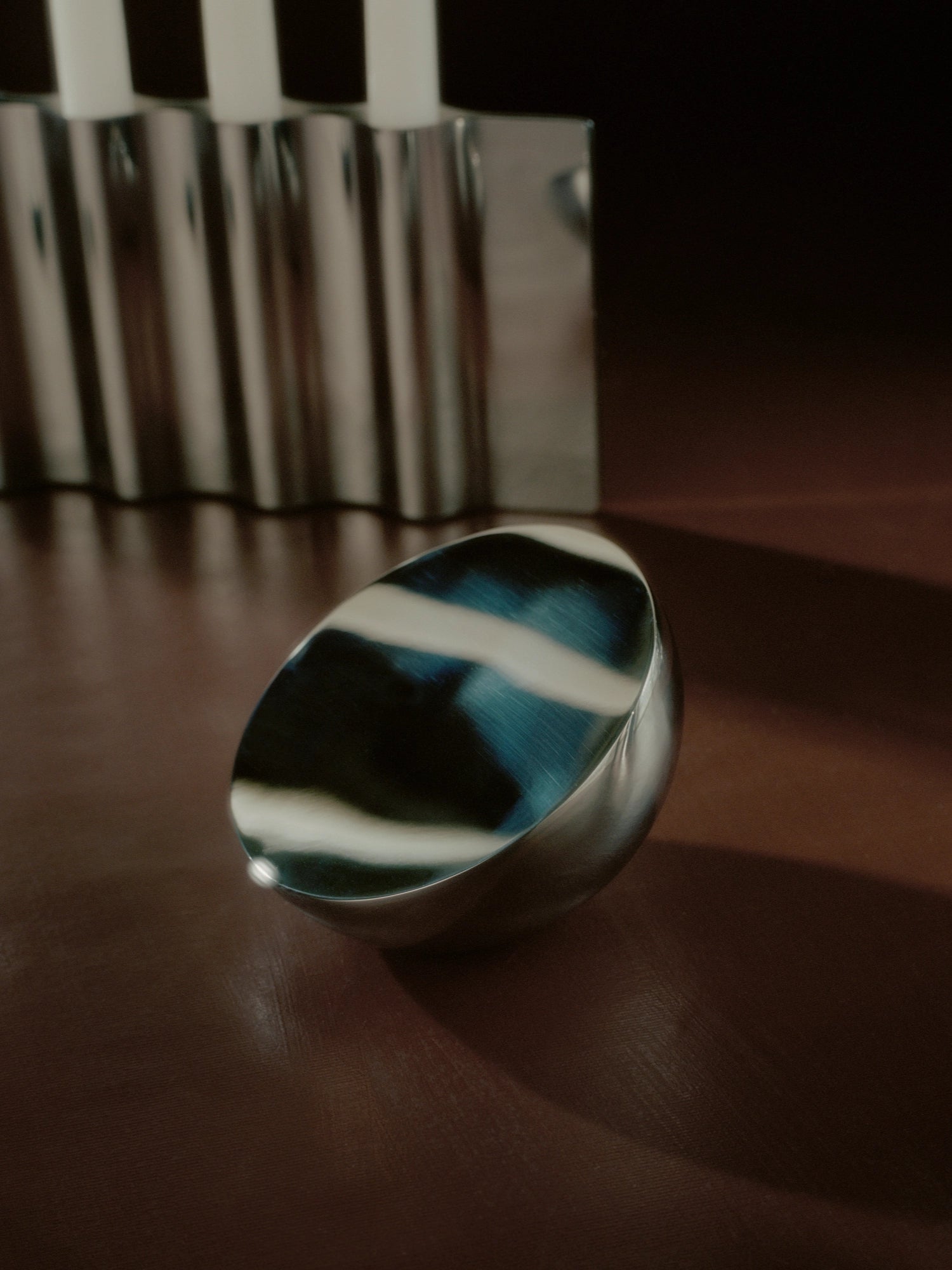 New Works Aura Table Mirror, a polished silver-tone metal semi-spherical decorative object with a reflective, deep blue and white abstract inner surface, displayed on a rich brown surface. A luxury accent piece.