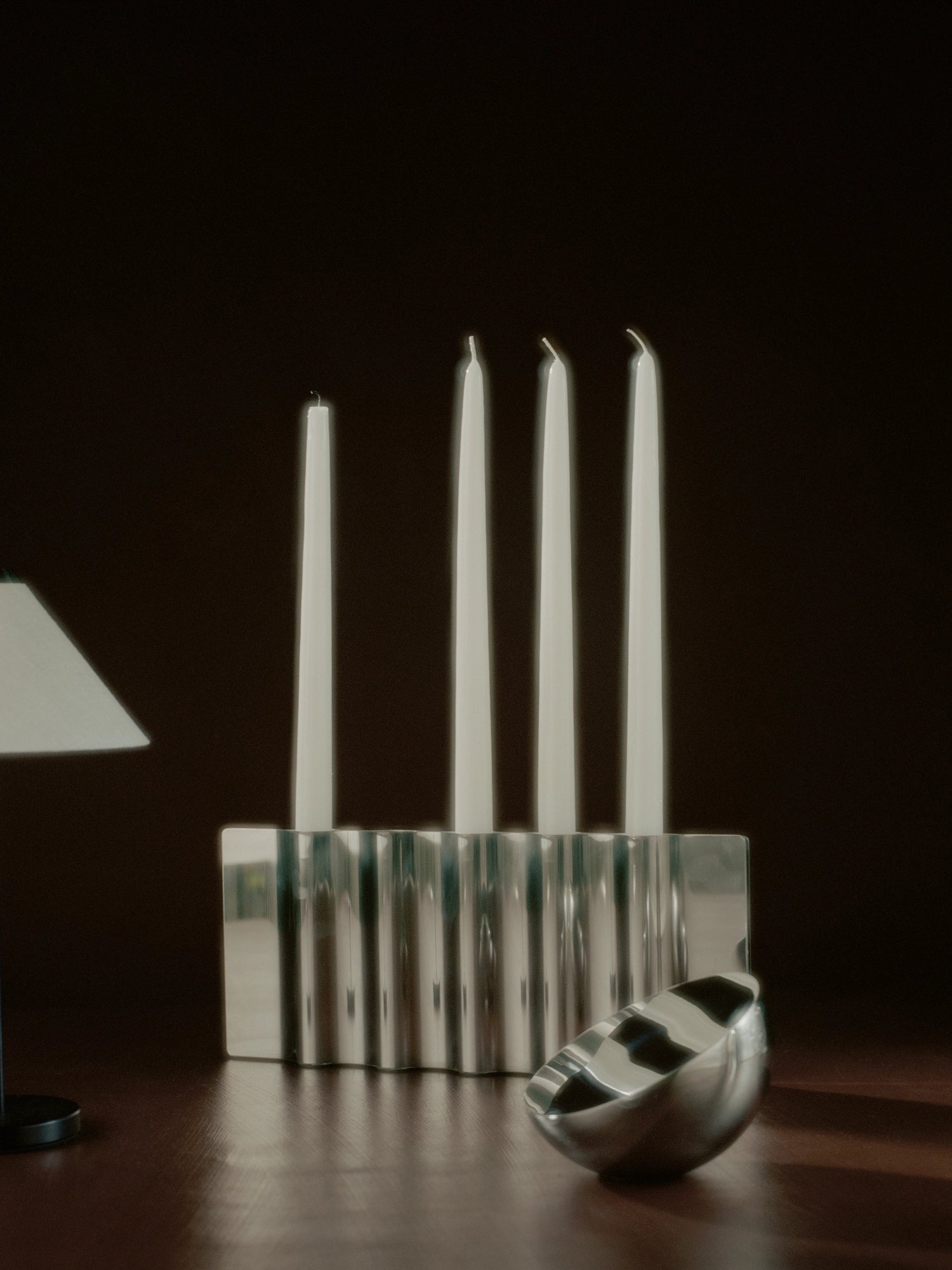 New Works Aura Table Mirror, showing its silver wavy, ribbed aluminum frame reflecting four white taper candles and a polished chrome bowl on a dark wood surface. A premium, modern decorative accent.