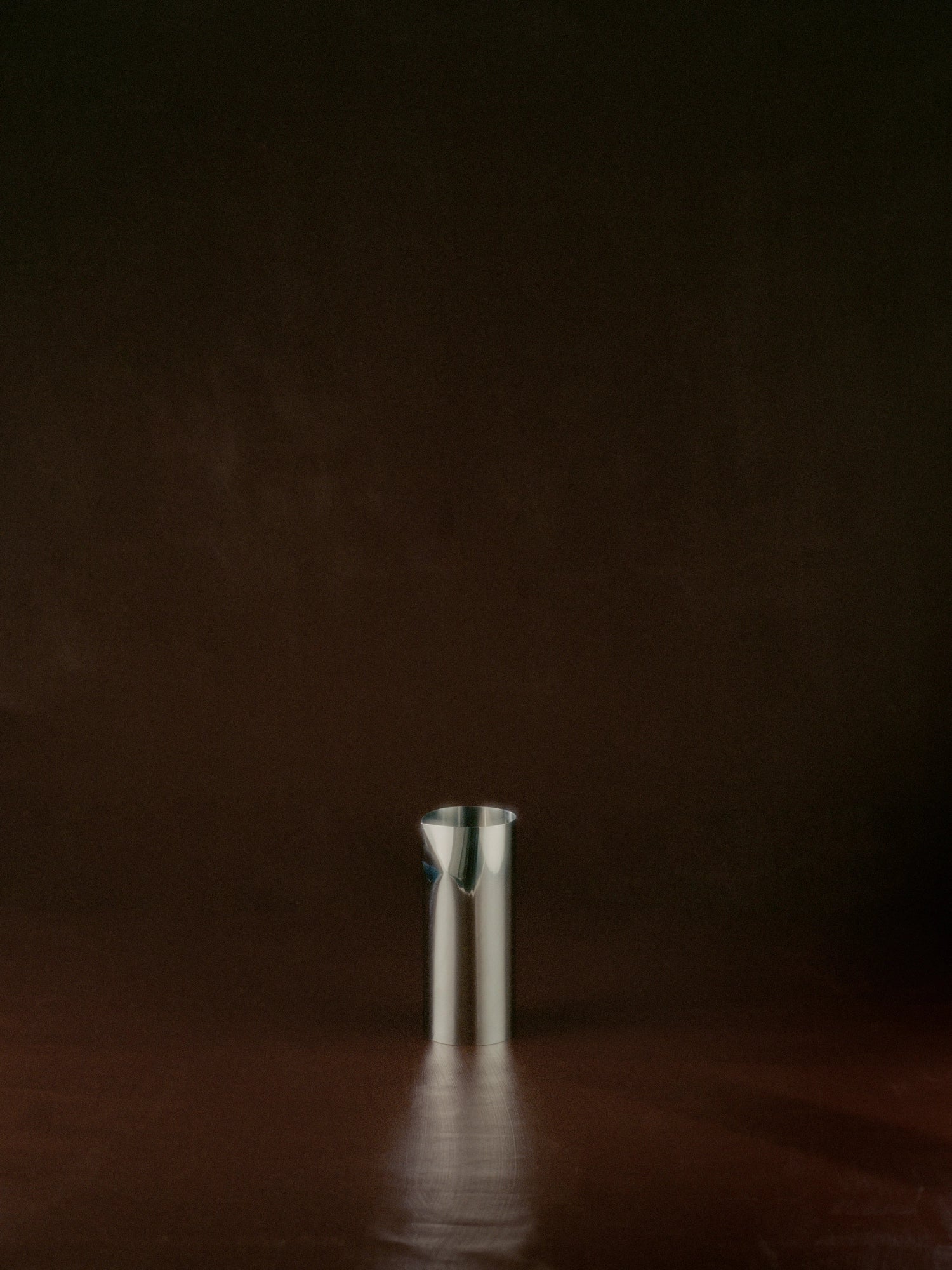 New Works Pleat Pitcher, a sleek polished silver steel beverage pitcher with a distinctive V-shaped spout, presented as a premium home good on a dark brown surface. Ideal for modern entertaining.