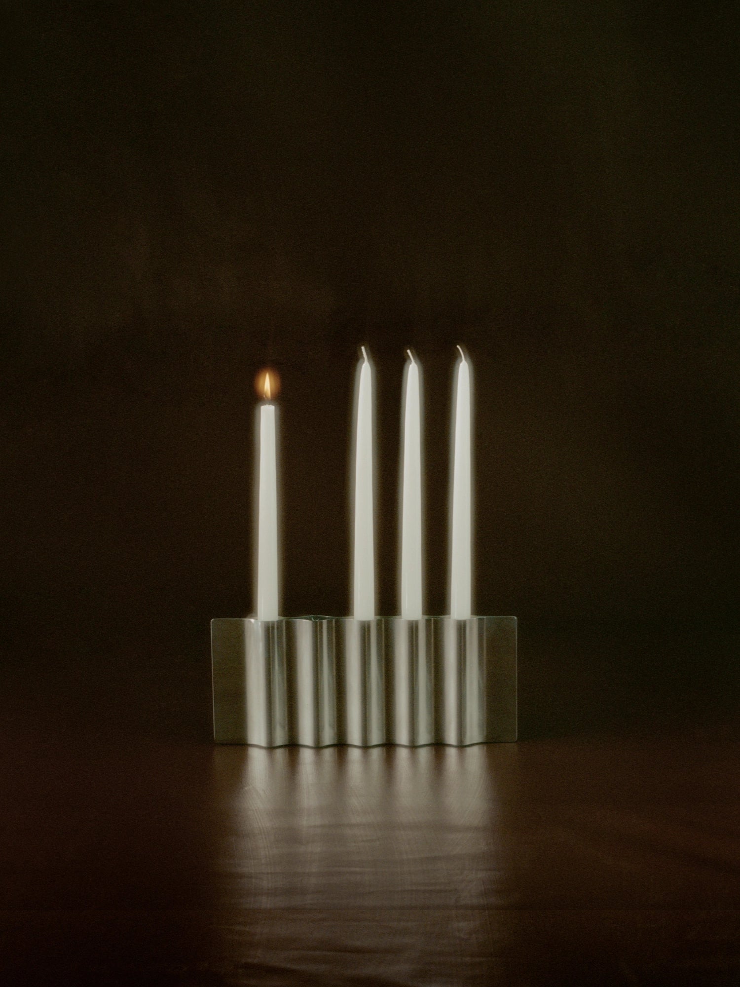 New Works Ripply Candle Holder, a modern, ripple-edged polished silver metal candle holder with four white taper candles, one glowing, displayed on a dark wooden surface. A luxurious design accent.