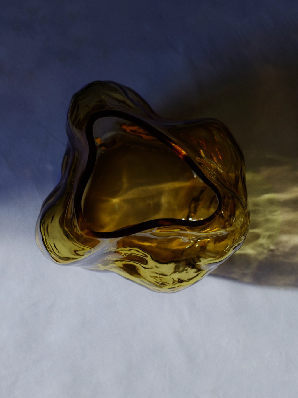 New Works A STUDY 001 / Blæhr Vase, a unique irregularly shaped amber-brown thick glass vase with rippling textures, displayed from an aerial view on a softly lit surface with shadows. A luxury statement piece for any modern interior.