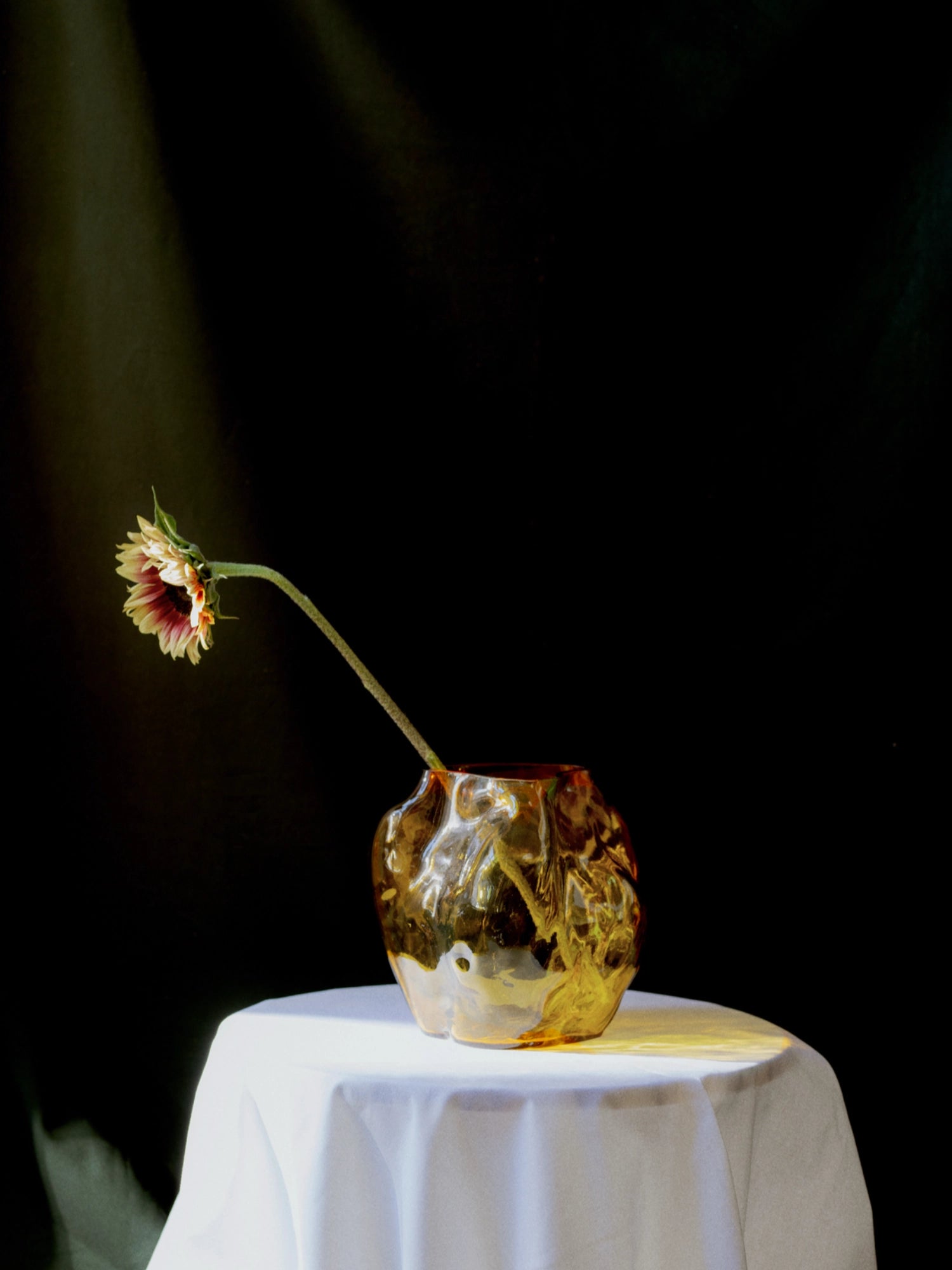 New Works A STUDY 001 / Blæhr Vase, a premium amber-colored sculpted glass artisan vase, with a subtle flower, displayed on a white tablecloth. Ideal for accentuating modern home decor.