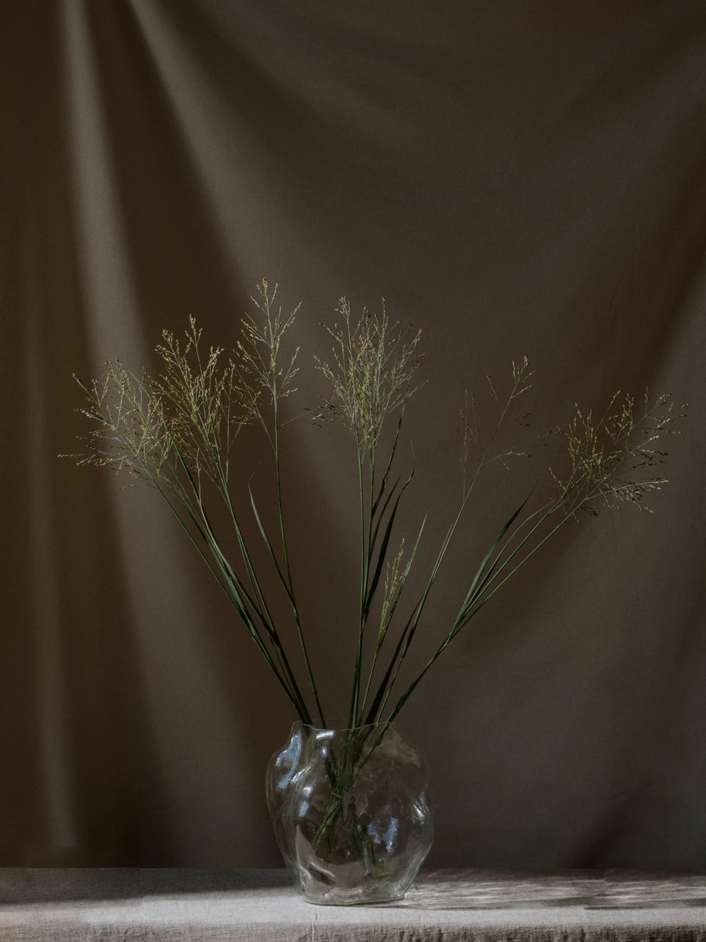 New Works A STUDY 001 / Blæhr Vase, a premium clear glass vase with an organic, wavy texture, holds a simple arrangement of wispy green grasses on a light linen tablecloth. Optimal for minimalist decor.
