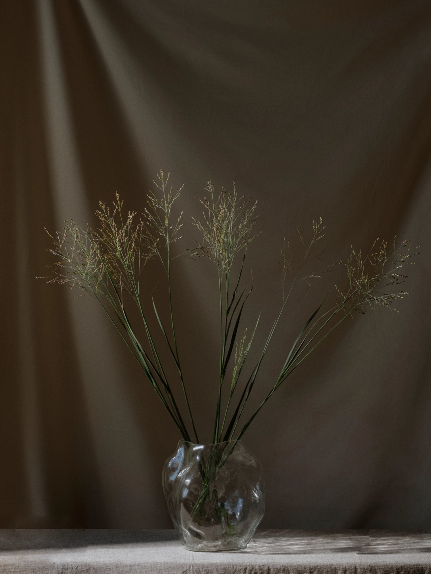 New Works A STUDY 001 / Blæhr Vase, a premium clear glass vase with an organic, wavy texture, holds a simple arrangement of wispy green grasses on a light linen tablecloth. Optimal for minimalist decor.
