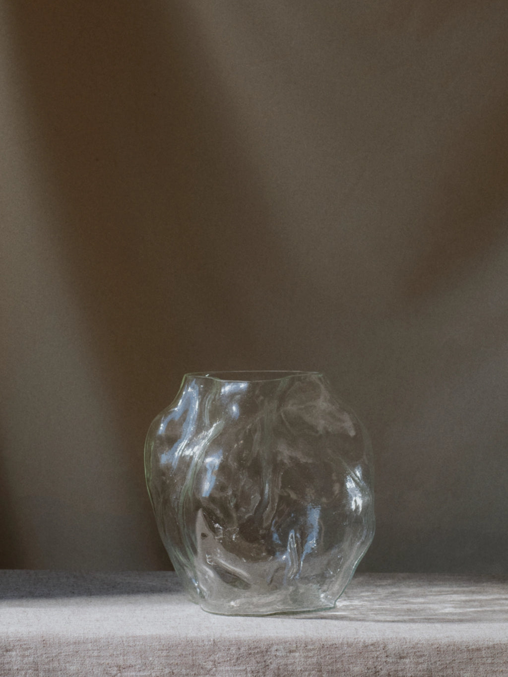 New Works A STUDY 001 / Blæhr Vase: a unique, clear glass vase with an organic, wavy, and uneven surface texture, showcased on a light linen-colored surface against a deep gray background. This luxury sculptural piece is ideal for modern home decor.