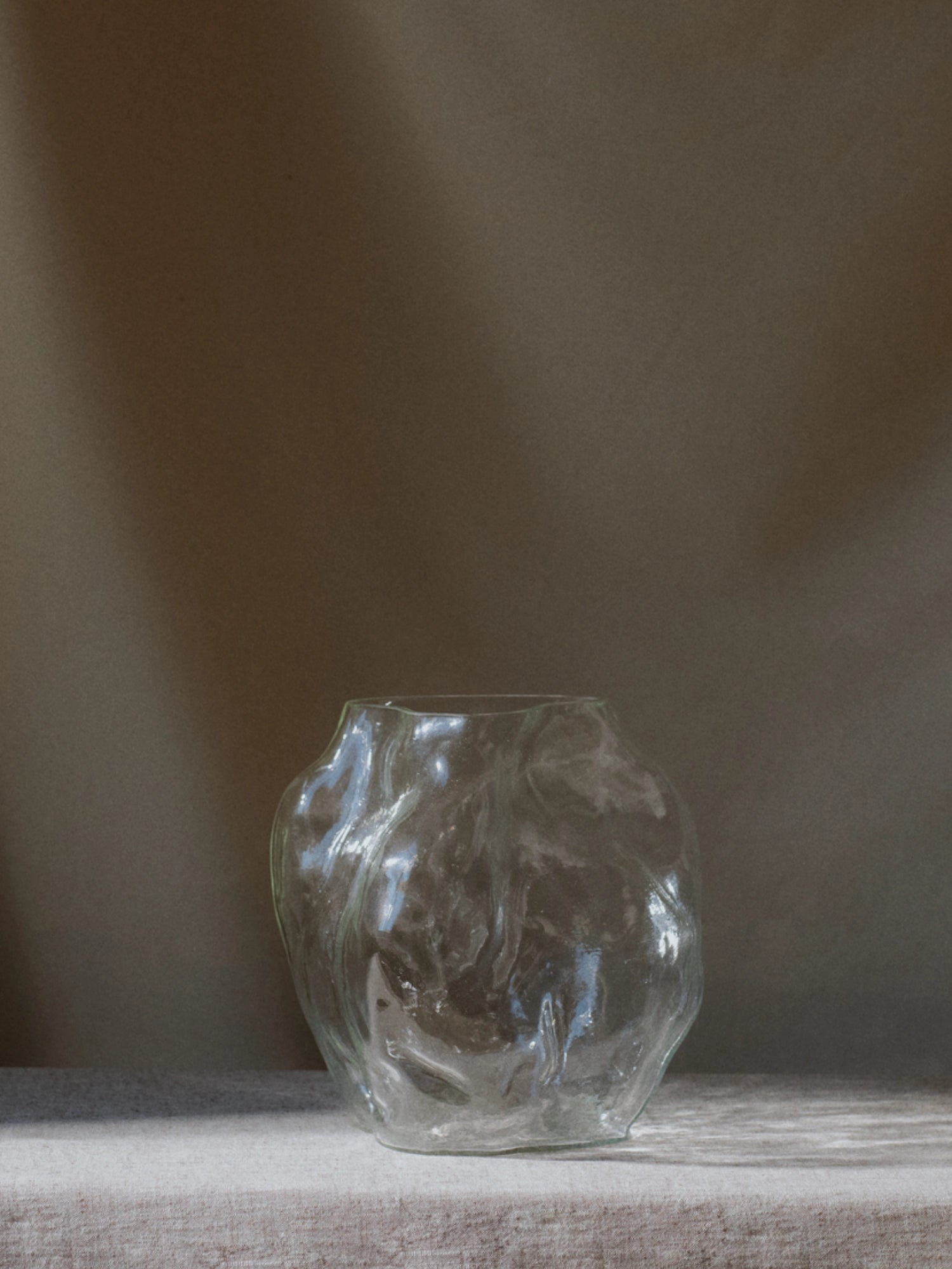 New Works A STUDY 001 / Blæhr Vase: a unique, clear glass vase with an organic, wavy, and uneven surface texture, showcased on a light linen-colored surface against a deep gray background. This luxury sculptural piece is ideal for modern home decor.