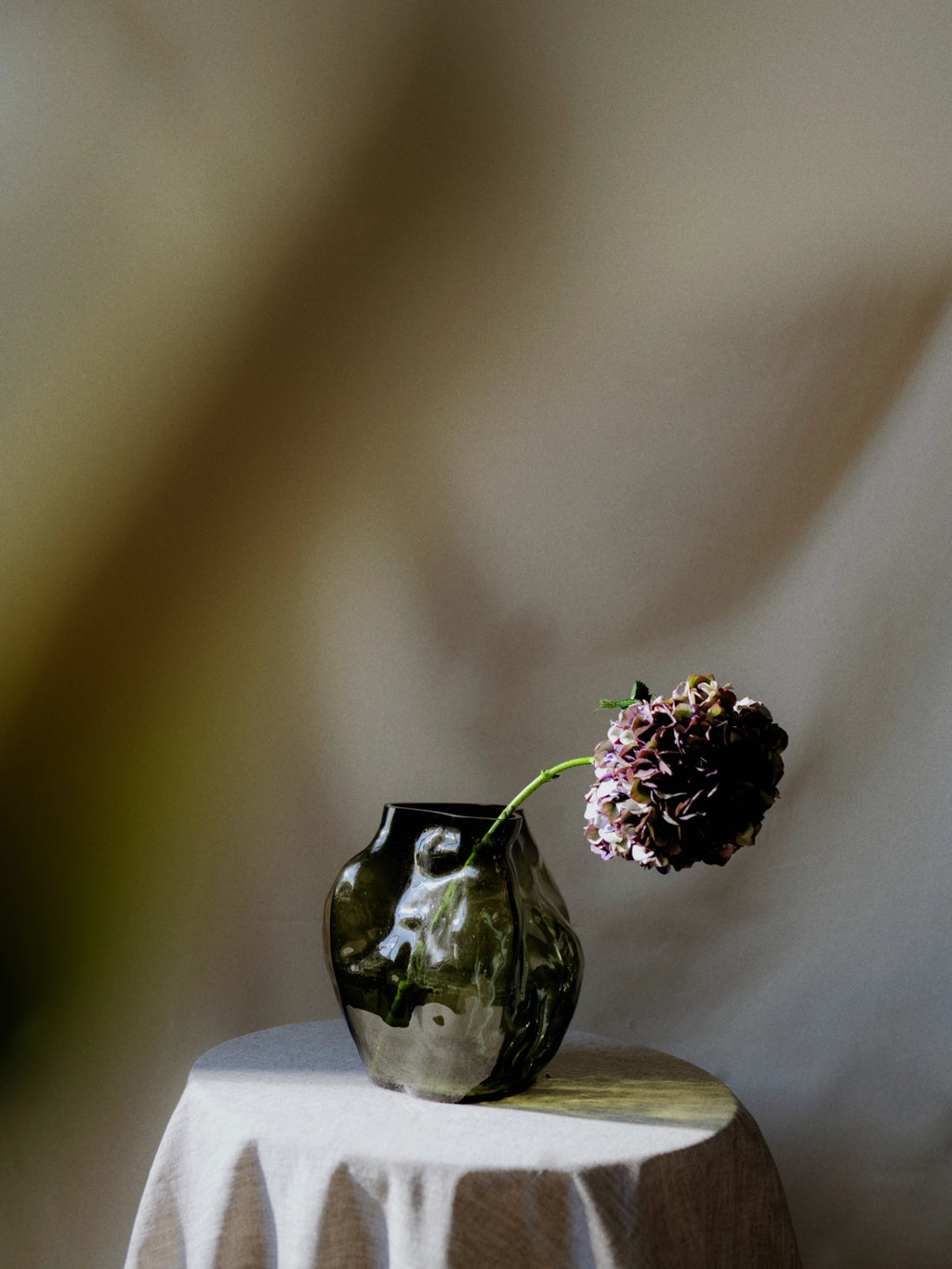 New Works A STUDY 001 / Blæhr Vase, a premium dark olive green organic-shaped glass vase with a glossy finish, holding a purple and green hydrangea bloom, displayed on a draped white linen table. Ideal for sophisticated home decor.