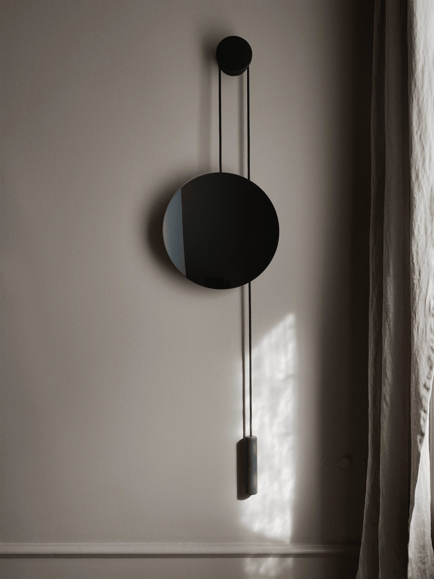 New Works Rise & Shine Wall Mirror: A minimalist round black mirror with an adjustable weight & pulley system in a lifestyle setting on a taupe wall, enhanced by natural light. Perfect for modern, stylish spaces.