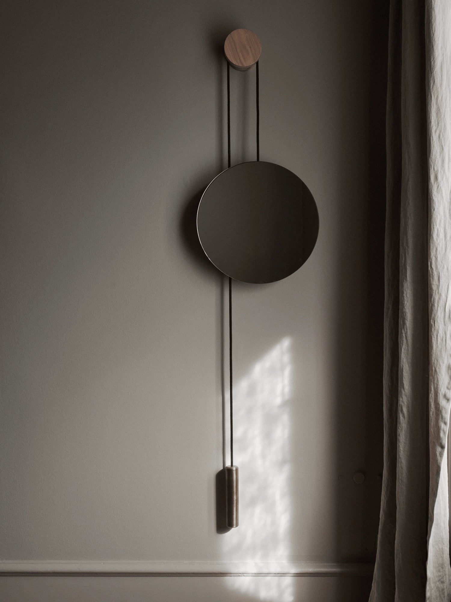New Works Rise & Shine Wall Mirror, a contemporary circular mirror with a polished brass frame, suspended by a black cord from a light wood disk and secured by a matching wood cylinder weight, displayed on a grey wall with a linen curtain. Ideal for Scandinavian modern decor.