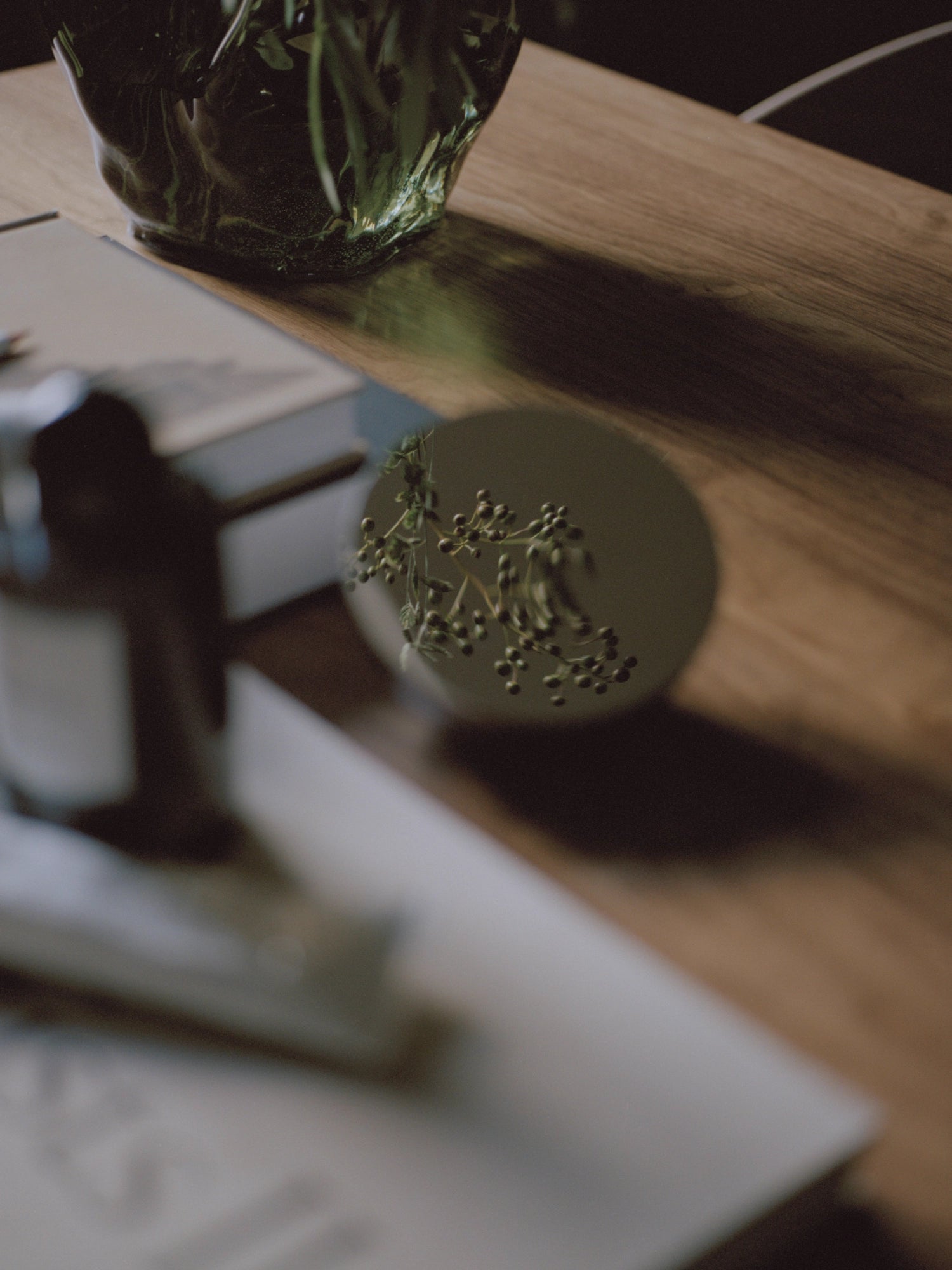 New Works Aura Table Mirror, circular, light gray, with reflective surface mirroring green plant details, styled on a warm brown wooden table next to a clear vase and book. Perfect for adding a premium, minimalist accent to any space.