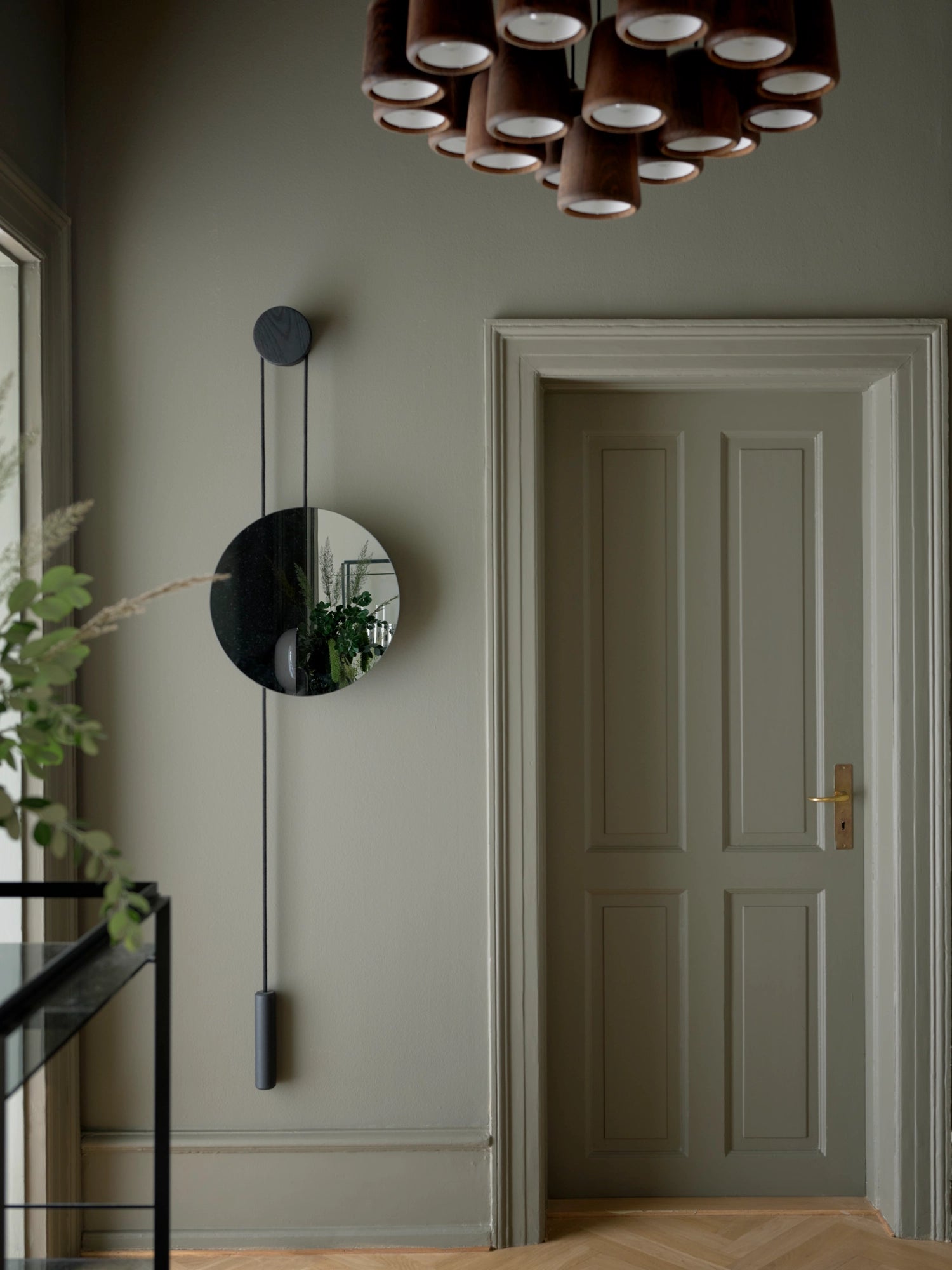 New Works Rise & Shine Wall Mirror; a stylish circular mirror with a black wooden support and counterweight, depicted in a modern entryway with a light olive-green wall. This luxury mirror adds a unique, functional design.