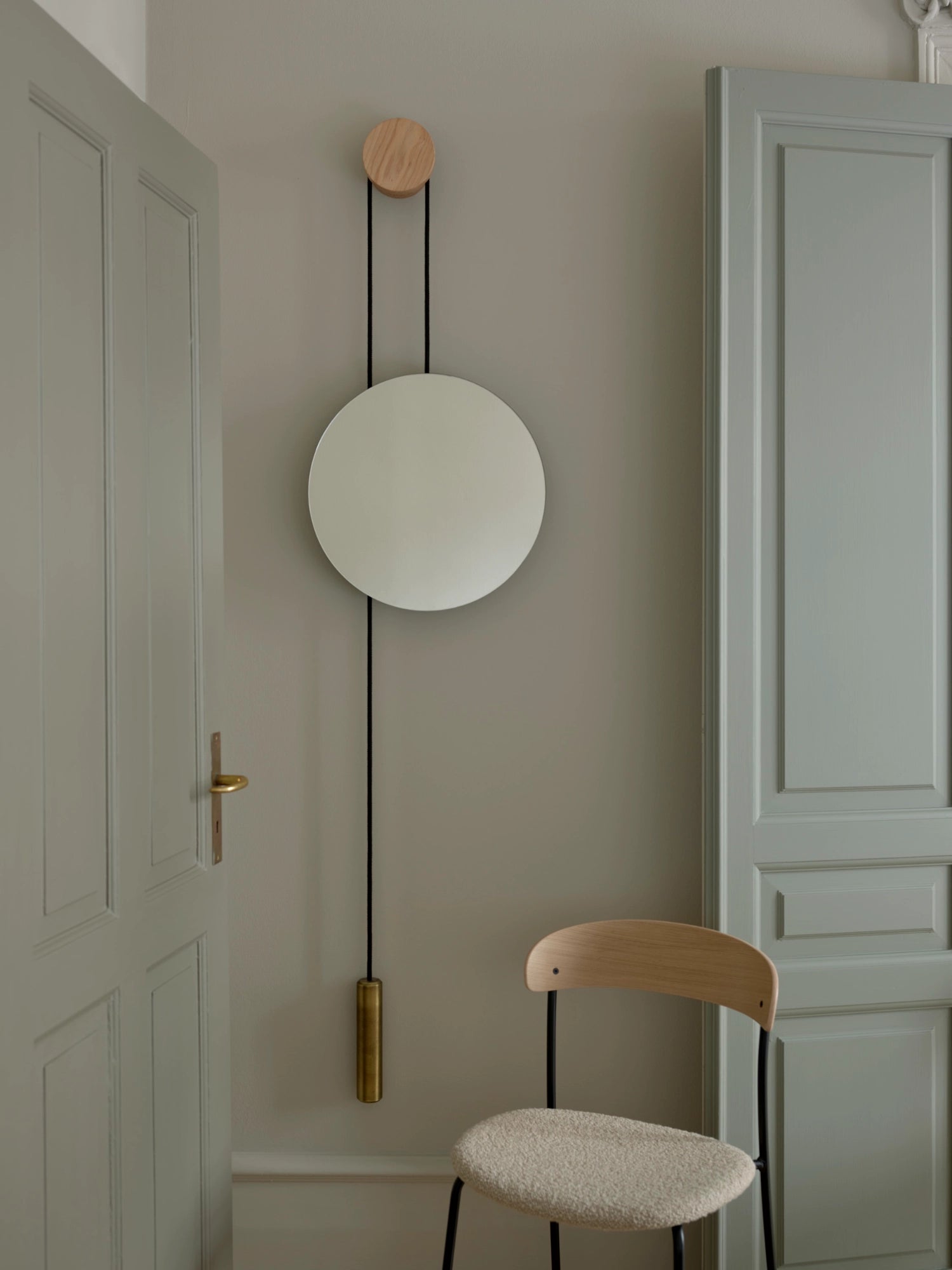 New Works Rise & Shine Wall Mirror, a luxurious round mirror with light oak mounting peg, black cord, and brass weight, displayed in a modern hallway. Perfect for contemporary interiors.