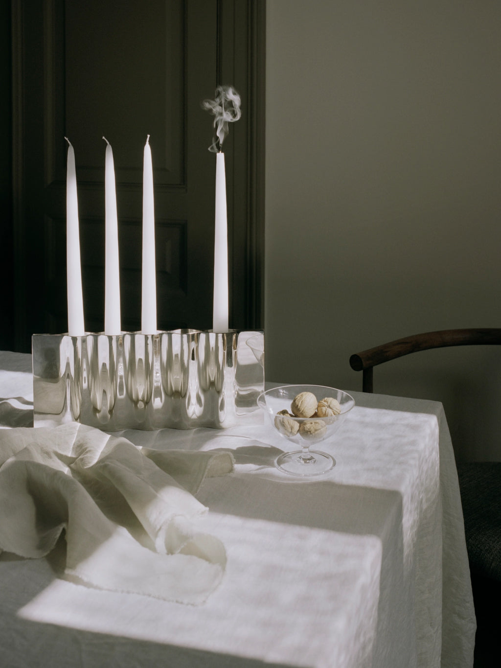 New Works Ripply Candle Holder: Elegant silver-toned metal holder with a distinctive rippled surface, displaying four white taper candles on a white tablecloth. This luxury centerpiece is ideal for sophisticated dining.