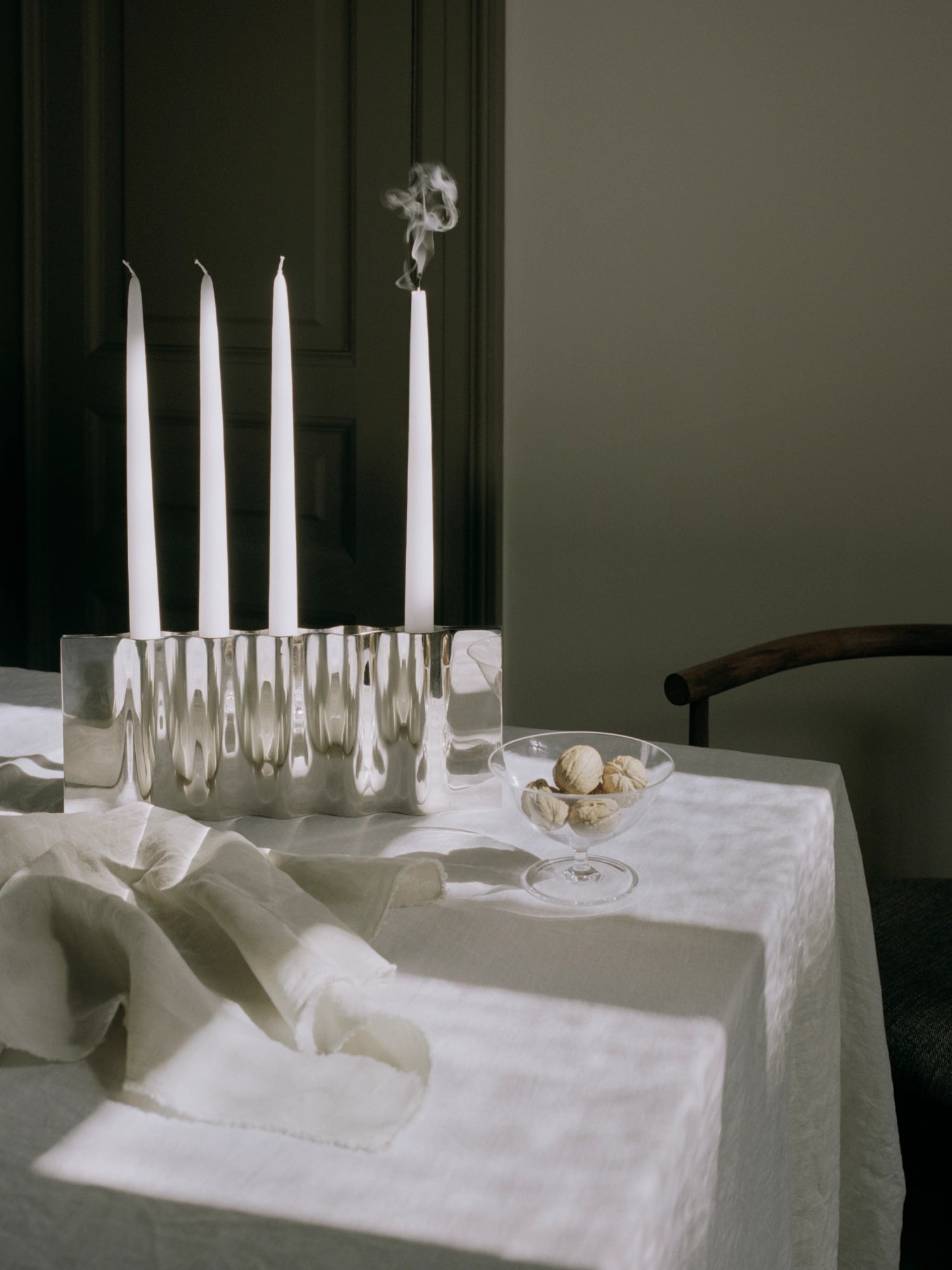 New Works Ripply Candle Holder: Elegant silver-toned metal holder with a distinctive rippled surface, displaying four white taper candles on a white tablecloth. This luxury centerpiece is ideal for sophisticated dining.
