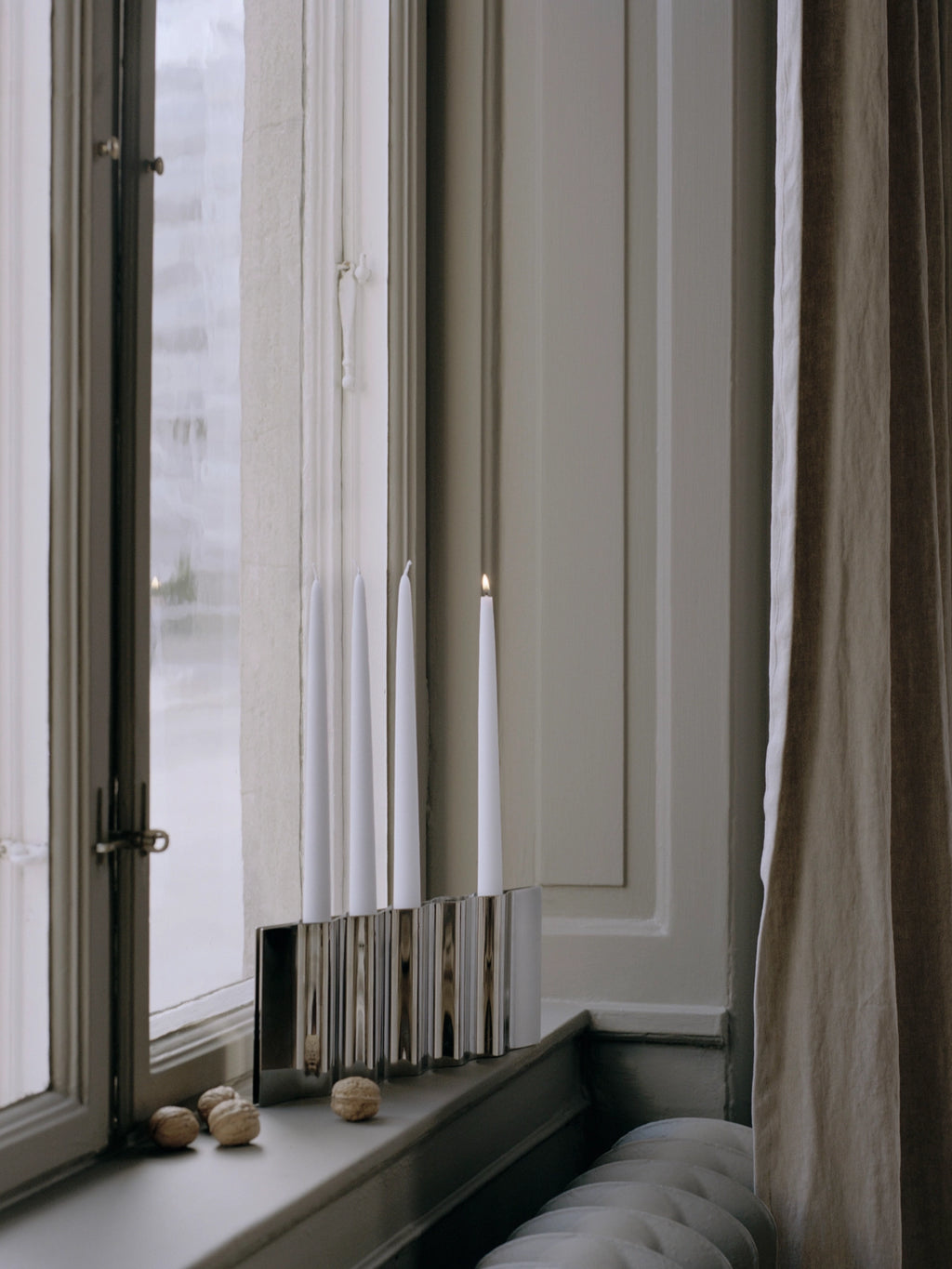 New Works Ripply Candle Holder, a modern silver-toned metallic candle holder with distinct vertical ridges, holding four tall white taper candles on a window sill next to walnuts, enhancing any contemporary decor.