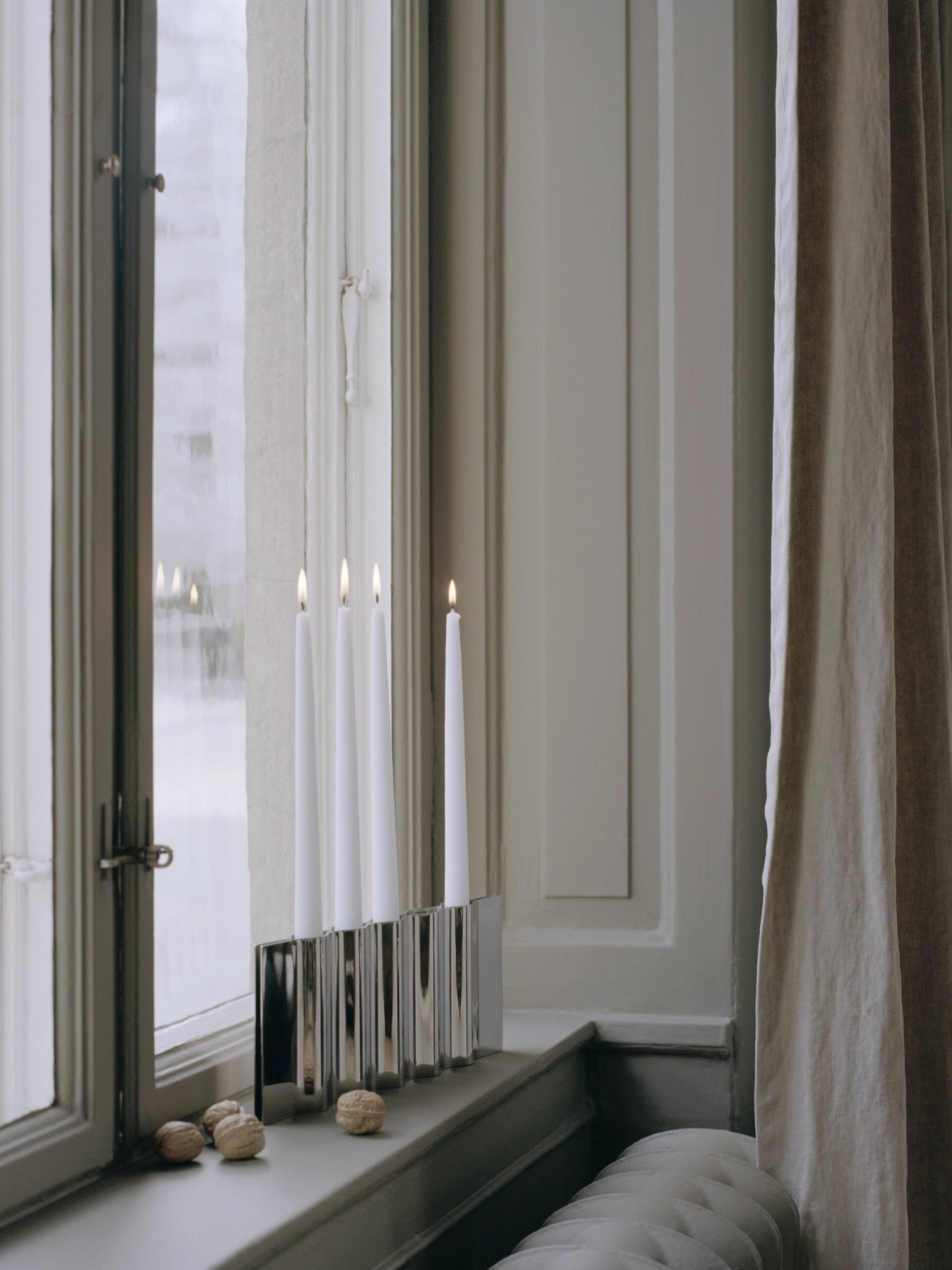 New Works Ripply Candle Holder: A premium polished silver metal holder with a distinctive ridged, ripple design, holding four lit white taper candles, displayed elegantly on a window sill with walnuts. Ideal for adding a luxurious touch to any interior.