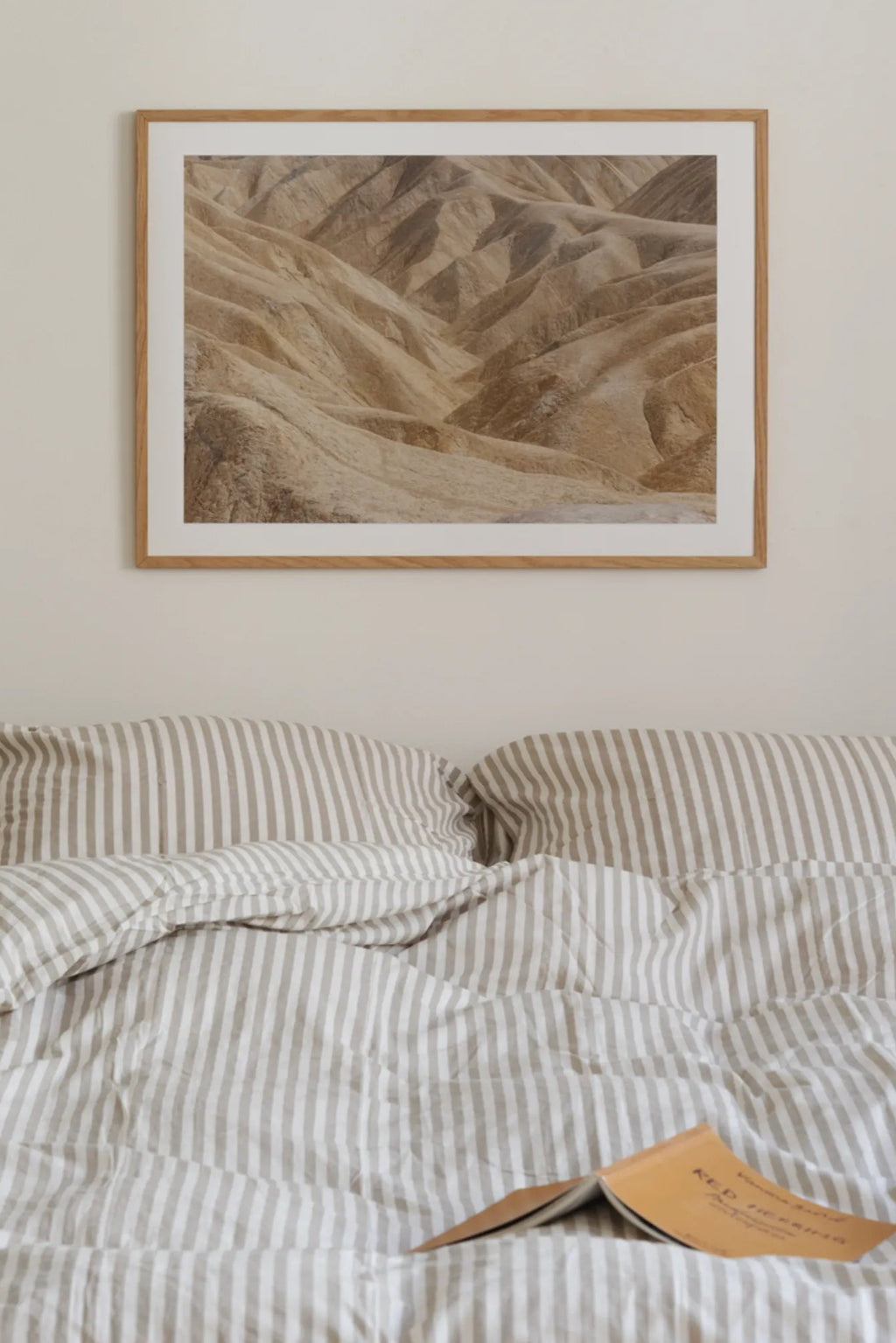 The Poster Club Perpetual, a captivating nature photography art print featuring a serene desert landscape with undulating sand dunes and natural earthy tones of beige, sand, and light brown. This minimalist abstract landscape poster is presented with a crisp white mat and a light natural oak frame. Perfect for creating a calming atmosphere in a Scandinavian bedroom, organic modern living room, or contemporary home office.