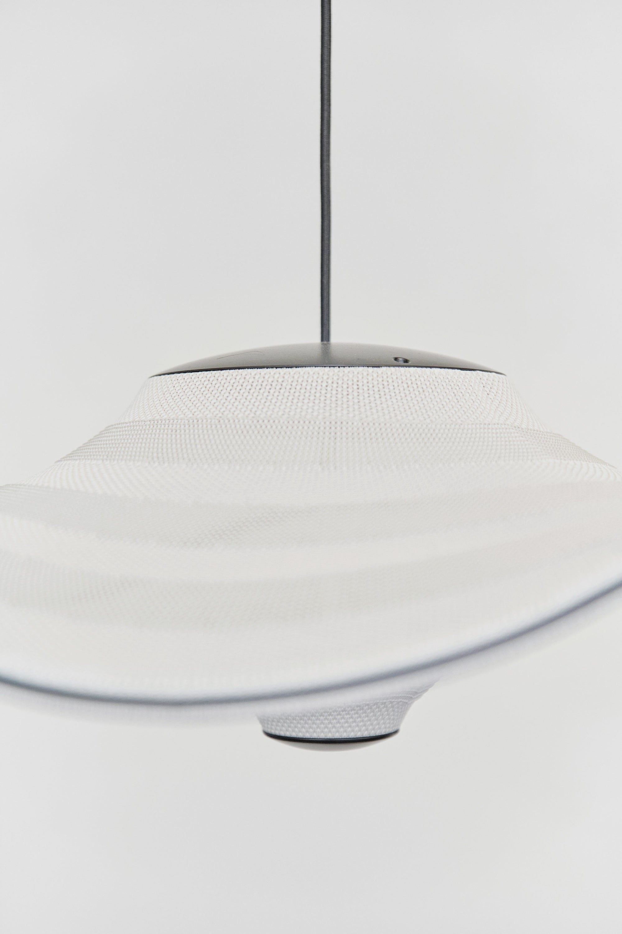 Made by Hand Flying Ø58 Pendant Lamp, a modern minimalist ceiling light featuring an off-white knitted textile shade with a subtly wavy, organic silhouette and a matte black metal top canopy and bottom accent. This Scandinavian pendant lamp provides soft, diffused light, perfect for contemporary living rooms, dining rooms, or bedrooms seeking elegant, organic modern lighting.