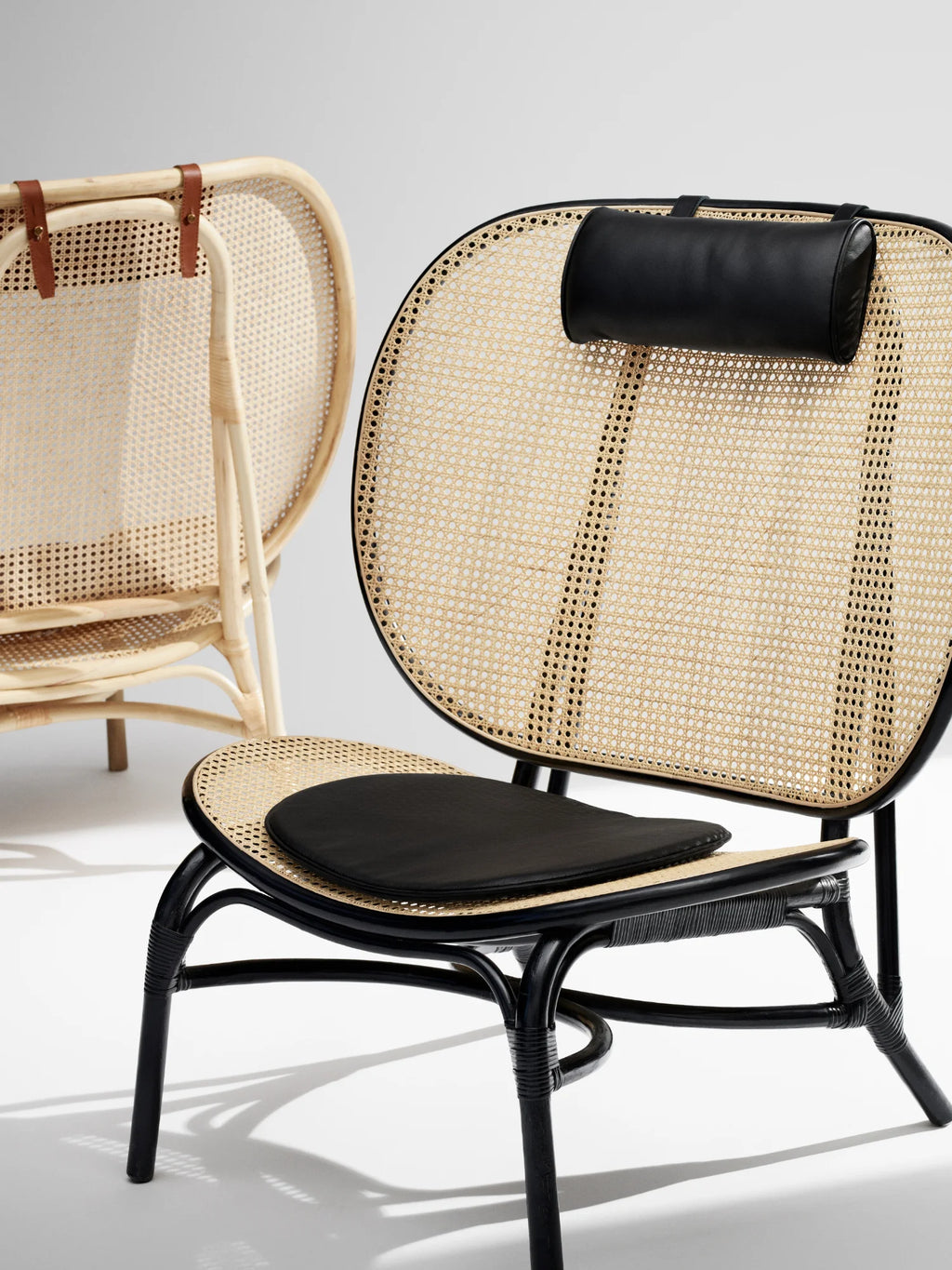 NORR11 Nomad Chair, featuring a rounded cane back and seat with a black leather cushion and headrest, and an elegant black lacquered rattan frame. This stylish lounge chair is perfect for adding a touch of modern comfort to any sophisticated living space.