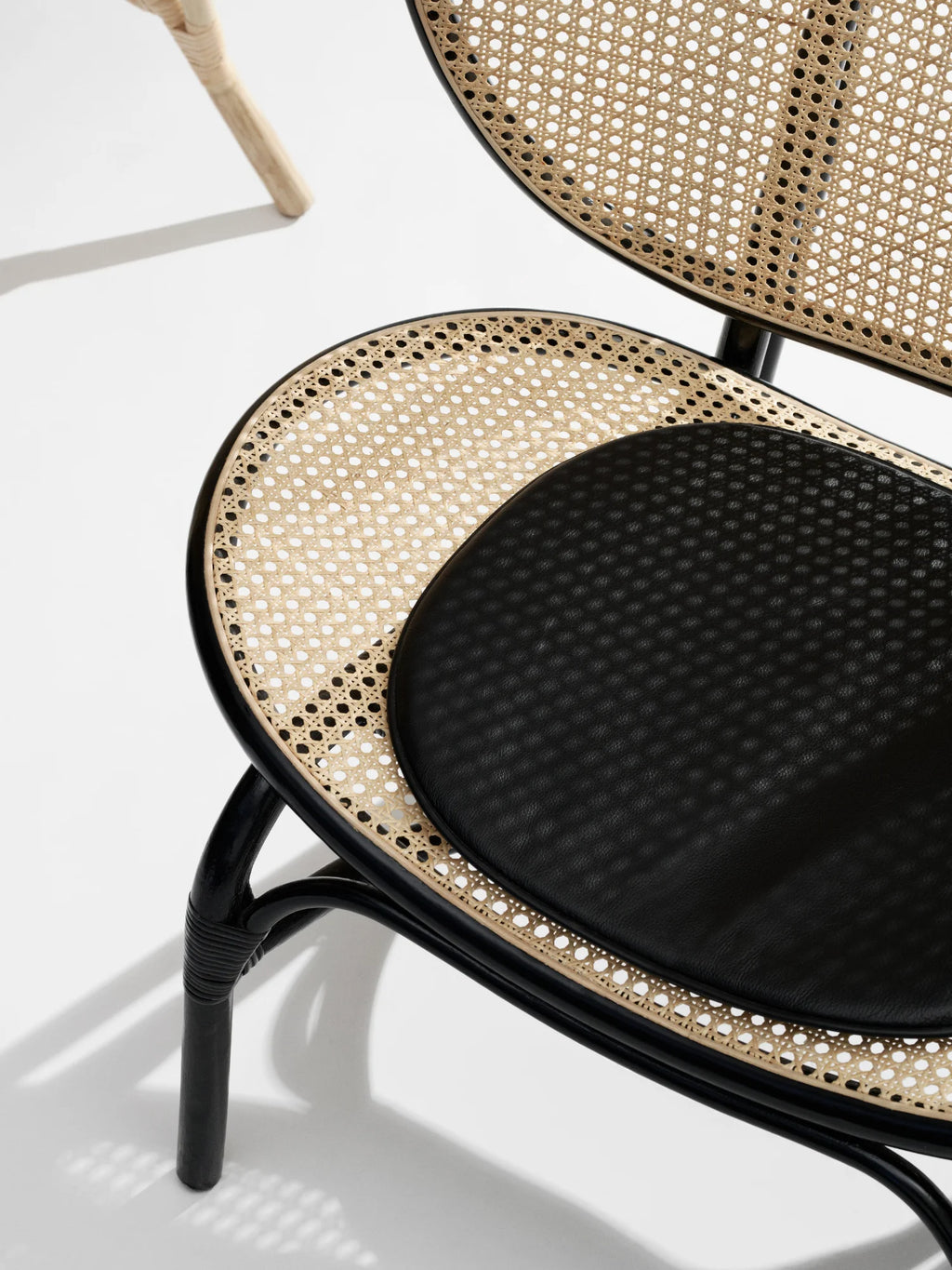 NORR11 Nomad Chair, a modern lounge chair featuring a black curved frame, natural cane webbing seat and back, and a black upholstered cushion. This elegant, handcrafted chair is perfect for adding a stylish and durable touch to any living space for those who appreciate sophisticated design.