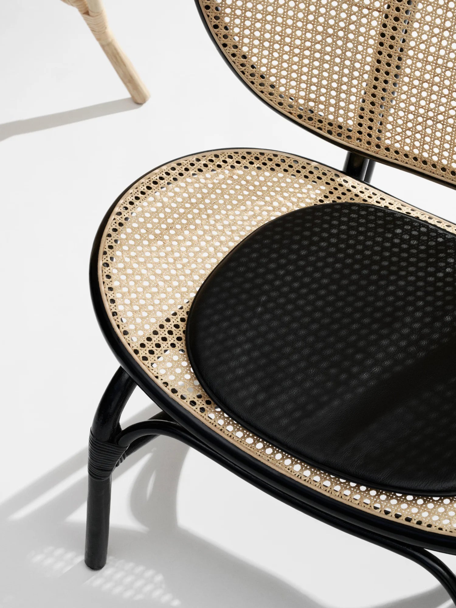 NORR11 Nomad Chair, a modern lounge chair featuring a black curved frame, natural cane webbing seat and back, and a black upholstered cushion. This elegant, handcrafted chair is perfect for adding a stylish and durable touch to any living space for those who appreciate sophisticated design.