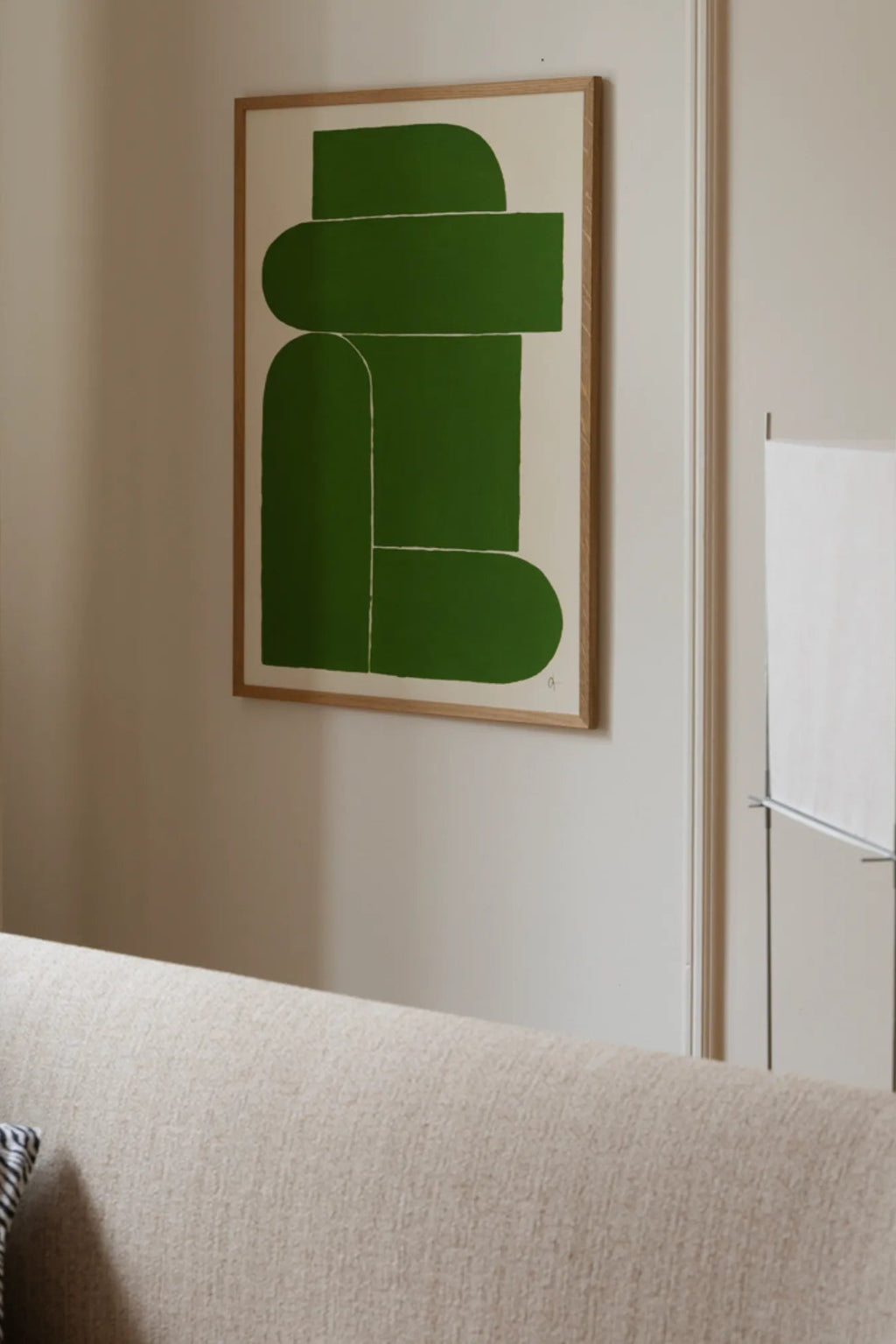 The Poster Club 9 pm, a minimalist abstract art print featuring bold deep green geometric shapes with soft rounded edges against a warm cream background, framed in a light natural oak frame. This contemporary wall art is perfect for Scandinavian living rooms, modern bedrooms, or gallery walls in organic modern interiors.