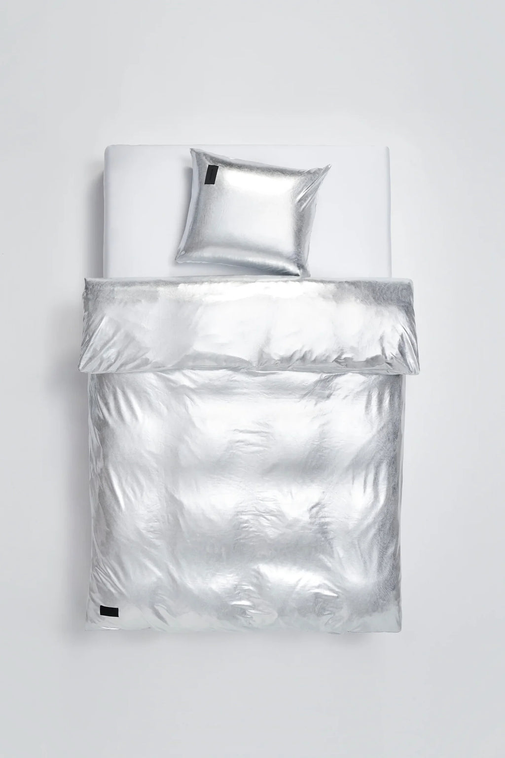 Magniberg Nude Metallic Jersey Pillow Case in a striking silver metallic finish, laid out flat on a white bed, showcasing its sleek, modern design and luxurious, reflective texture.