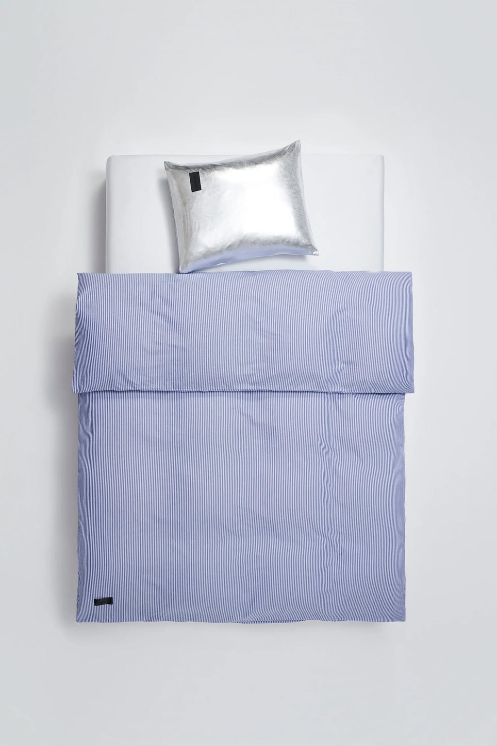 Magniberg Nude Metallic Jersey Pillow Case in a striking silver metallic finish, paired with a blue striped duvet cover, creating a unique and modern bedroom aesthetic, displayed on a white bed.