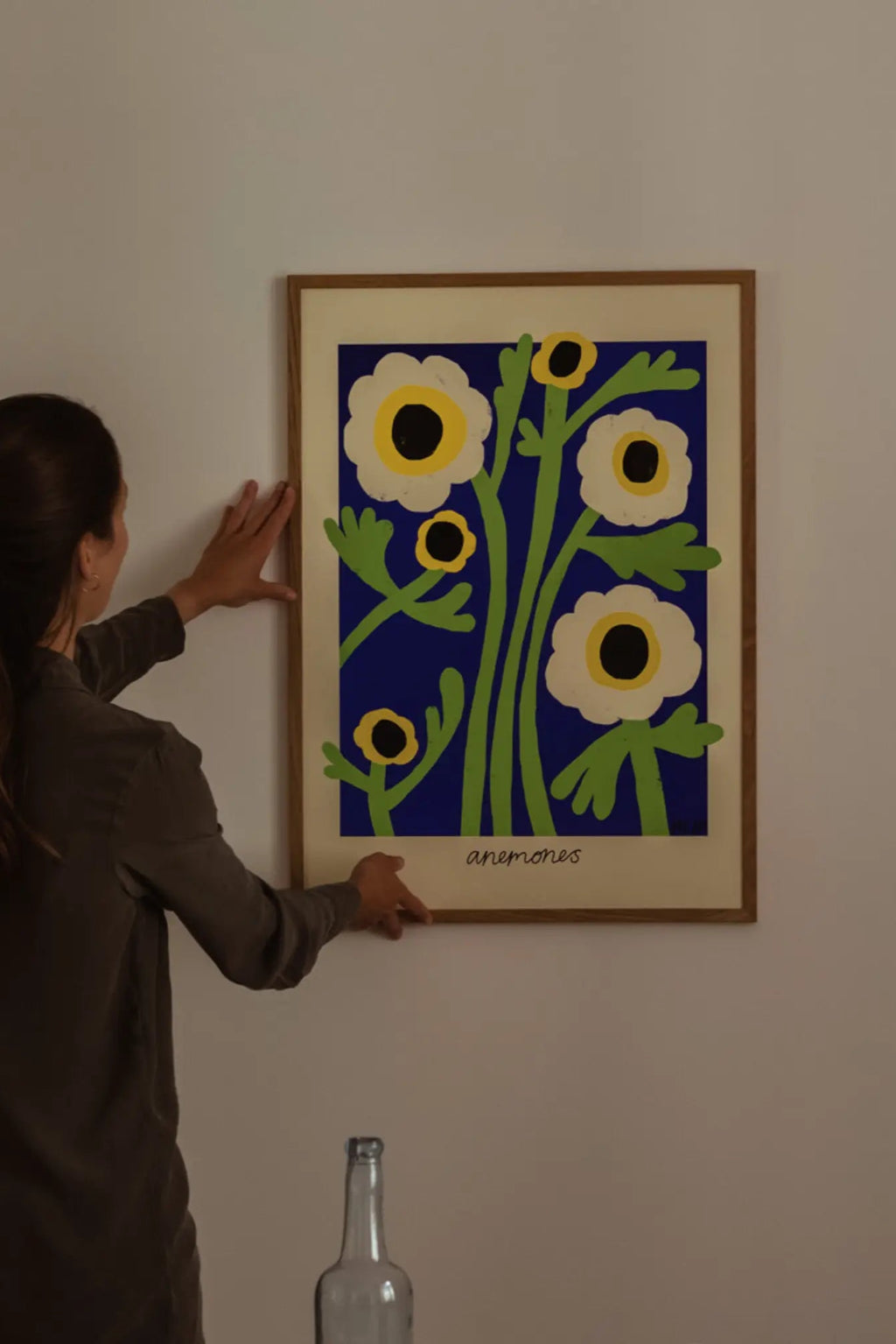The Poster Club Yellow Anemones, a vibrant modern floral art print featuring graphic white anemone flowers with yellow and black centers and lime green stems against a bold royal blue background, presented with a cream mat in a light natural oak frame. This contemporary botanical poster is ideal for adding a pop of color to Scandinavian living rooms, minimalist bedrooms, or a stylish gallery wall.