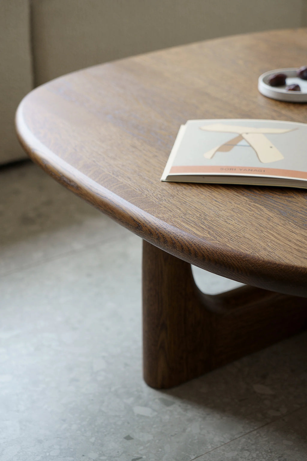 TAMO Olfo Coffee Table, a close-up of the premium dark brown wooden tabletop with beautiful grain detailing and elegantly curved edges, styled next to a book. Perfect for modern living spaces.