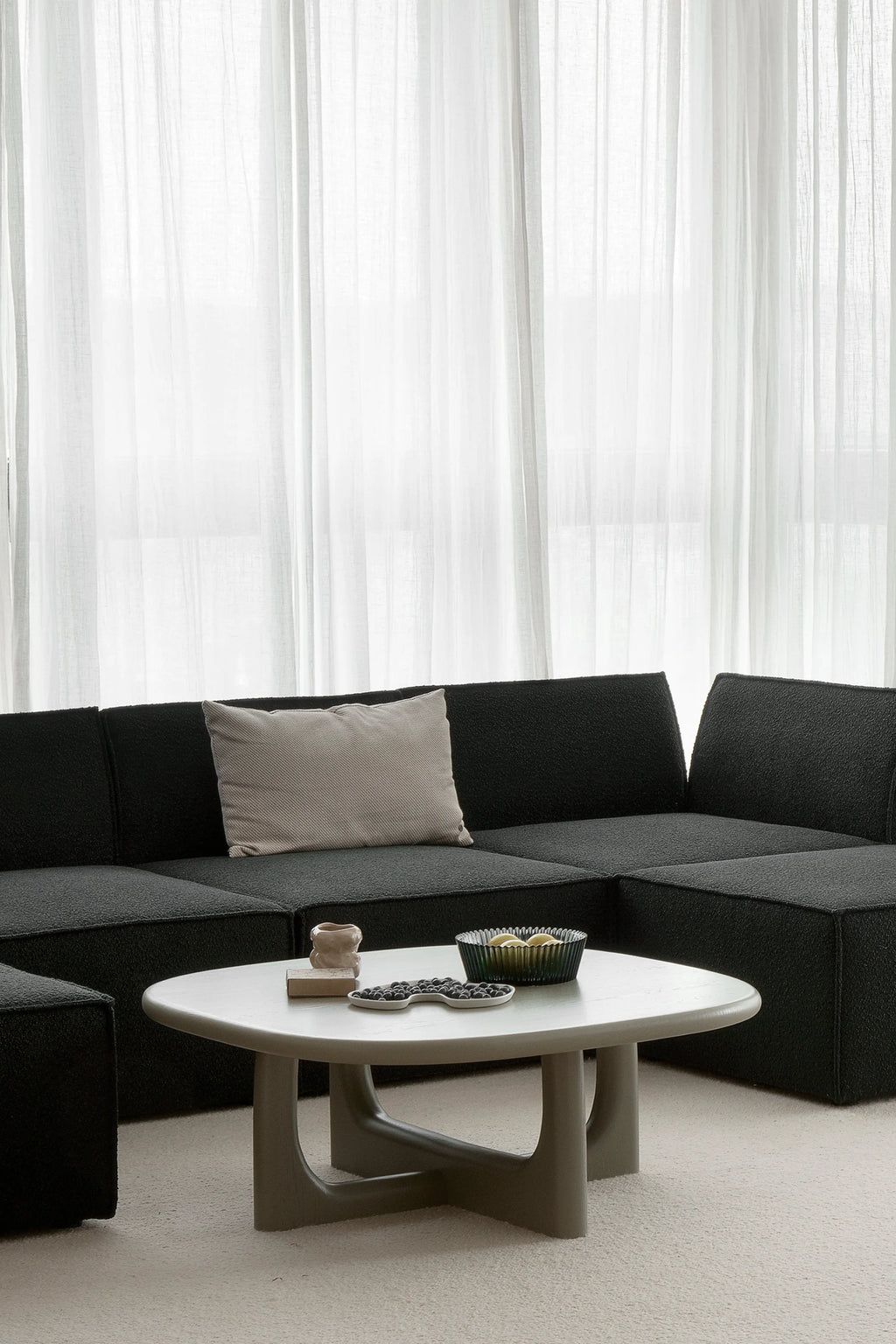 TAMO Olfo Coffee Table, a premium oval light grey wooden coffee table with unique cross-leg supports, next to a black sectional sofa. Ideal for modern living spaces.