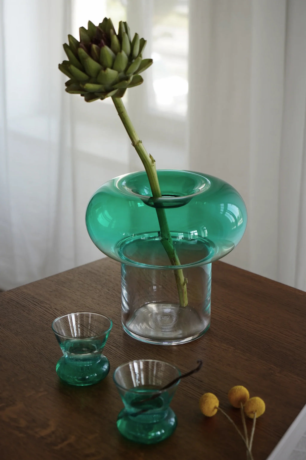 TAMO Orio Drinking Glass - Set of 2. Two small translucent hourglass-shaped glasses with deep teal bases on a light-brown wooden table beside a green glass vase and a vanilla bean stick. Ideal for unique beverage presentation.