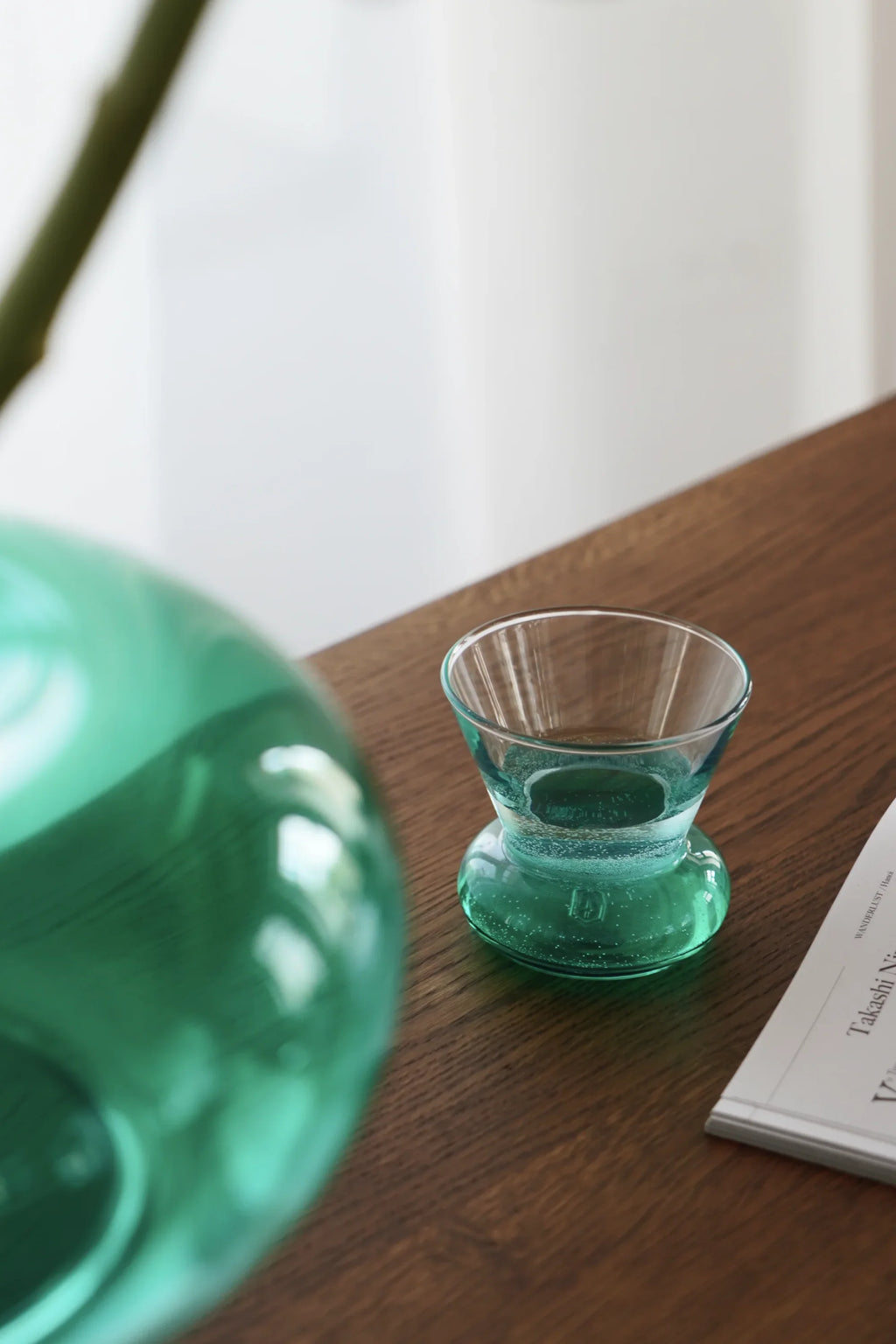 TAMO Orio Drinking Glass (Set of 2) with a unique hourglass shape, crafted from bubbly teal-tinted premium glass, displayed with water on a wooden table. Ideal for stylish beverage serving.