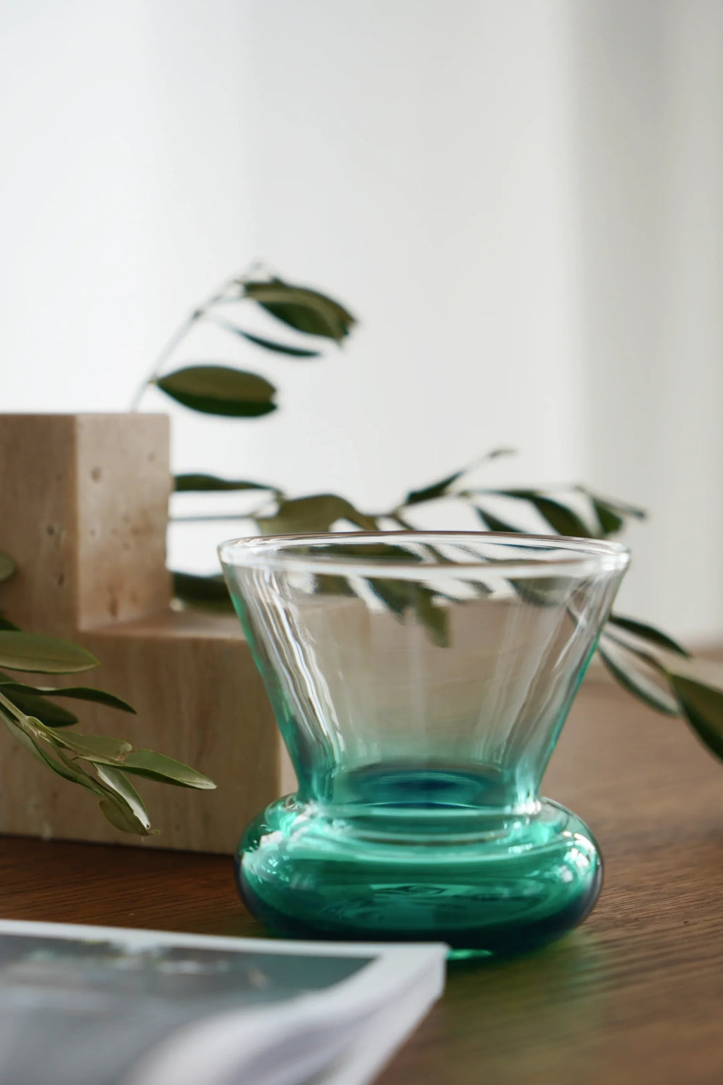 TAMO Orio Drinking Glass with a clear fanned top, graduating to a vibrant teal-blue rounded base, showcasing a single premium glass from the Set of 2 on a wooden surface with a book, marble blocks, and olive leaves. Perfect for elegant beverage serving.