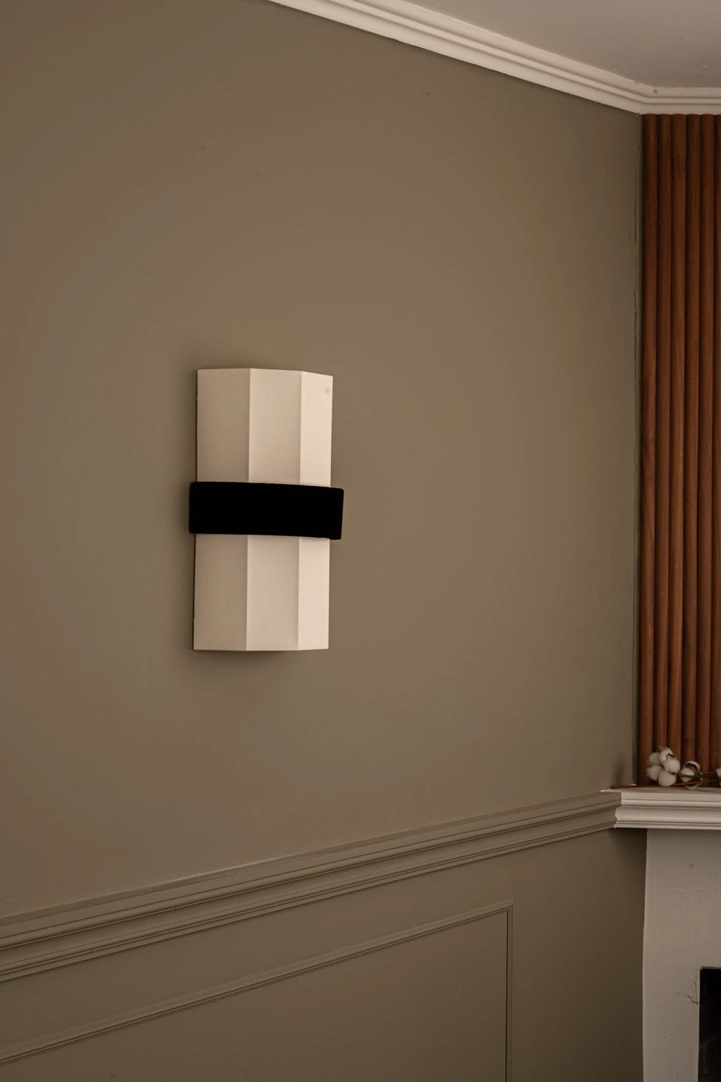 Simone & Marcel Okla Wall Light, a modern sculptural wall sconce featuring a cream-colored faceted ceramic body with a striking matte black velvet band wrapped around its center. This contemporary geometric wall light offers a minimalist design aesthetic, casting ambient light upwards and downwards from its textured ceramic surface. Ideal for sophisticated living rooms, bedrooms, or entryways seeking a unique designer lighting fixture.