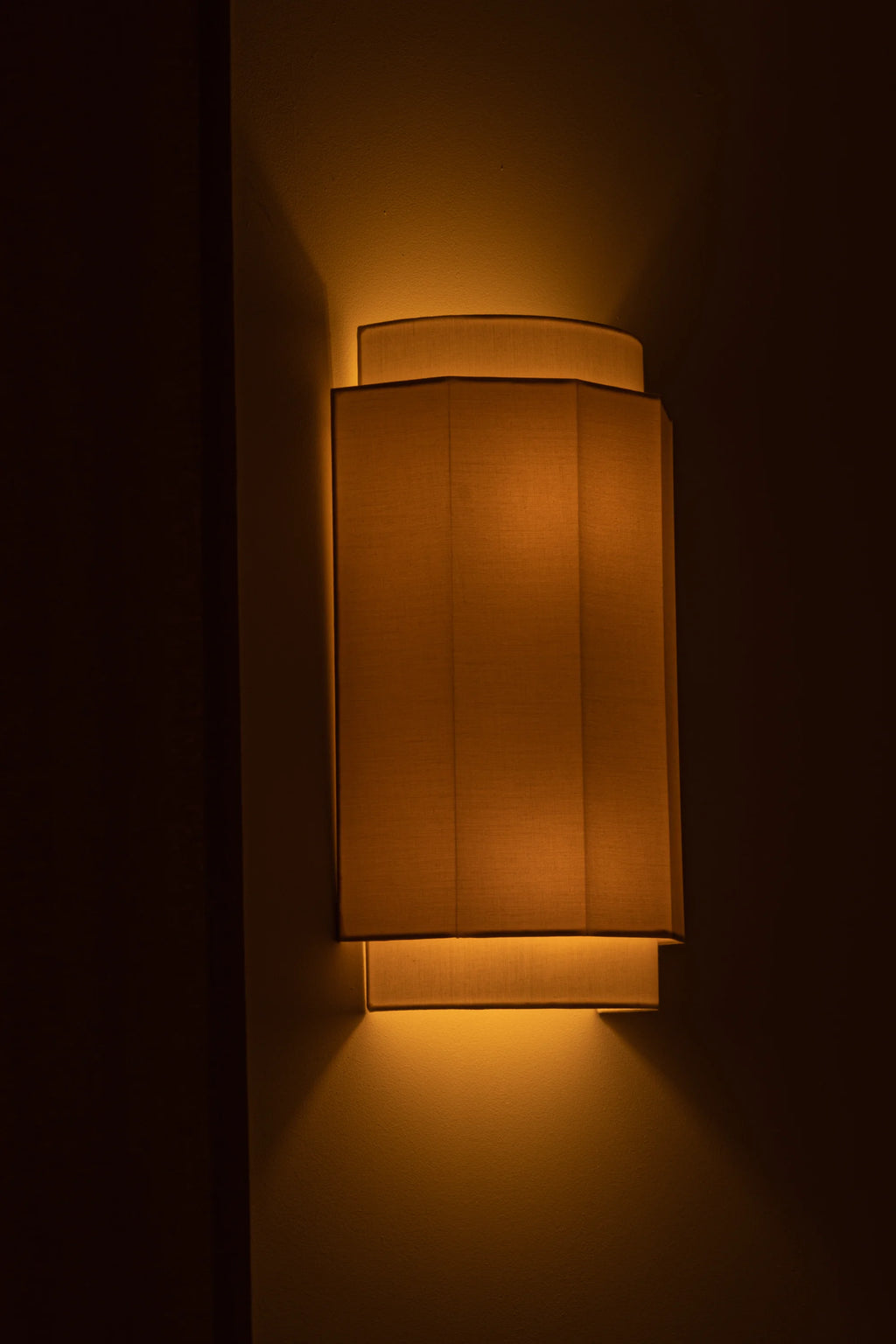 Simone & Marcel Okla Wall Light, a modern geometric wall sconce featuring a layered design with a rectangular, textured fabric shade that emits a warm amber glow. This contemporary wall light has elegant curved top and bottom plates, creating sophisticated up and down ambient lighting. Perfect for minimalist living rooms, cozy bedrooms, or stylish hallways.