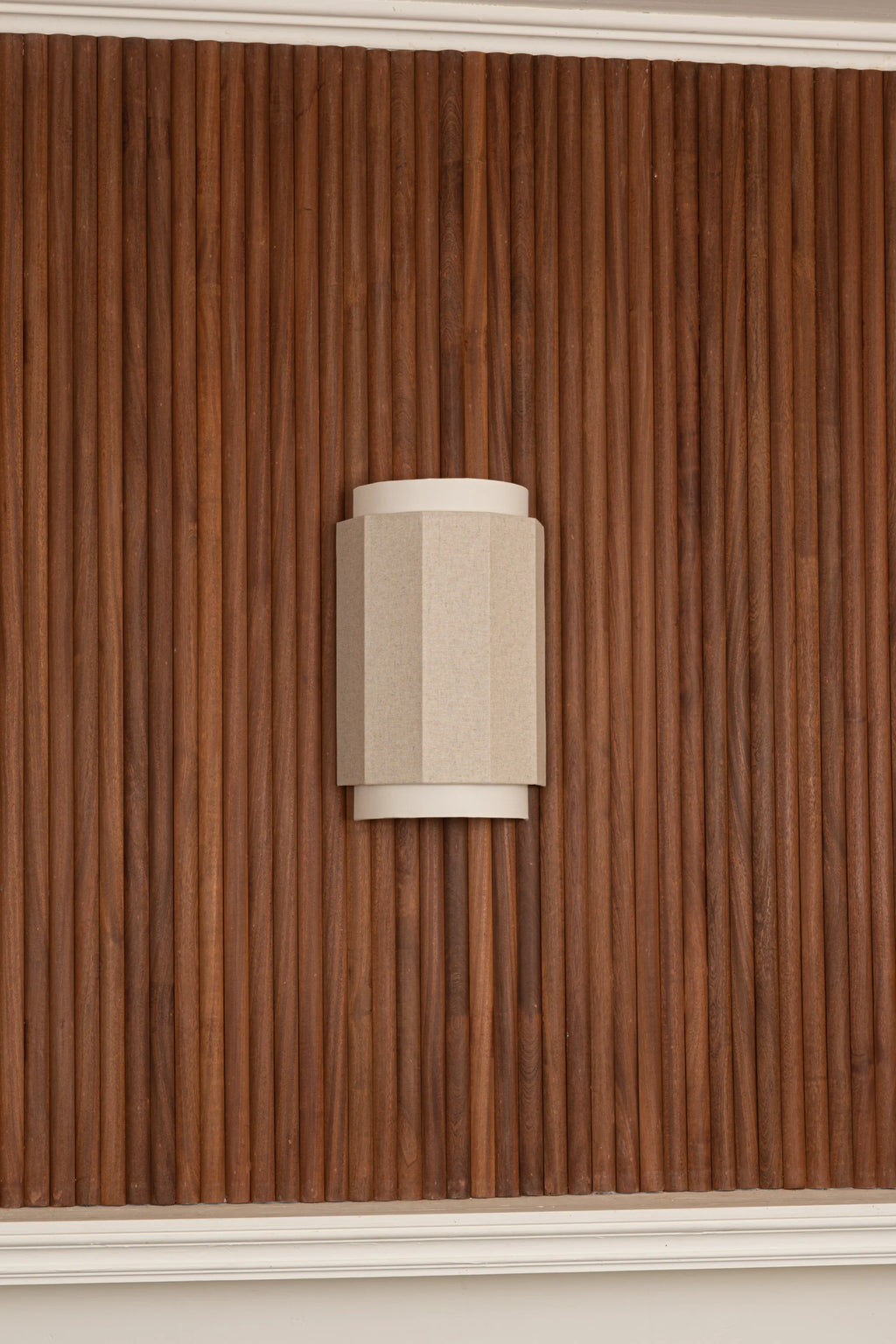 Simone & Marcel Okla Wall Light, a modern wall sconce featuring a geometric faceted shade crafted from natural beige linen fabric, resting on a smooth cream cylindrical base. This contemporary light fixture is designed for sophisticated interiors, perfect as an accent in minimalist living rooms, organic modern bedrooms, or elegant hallways, especially against rich wood paneling.
