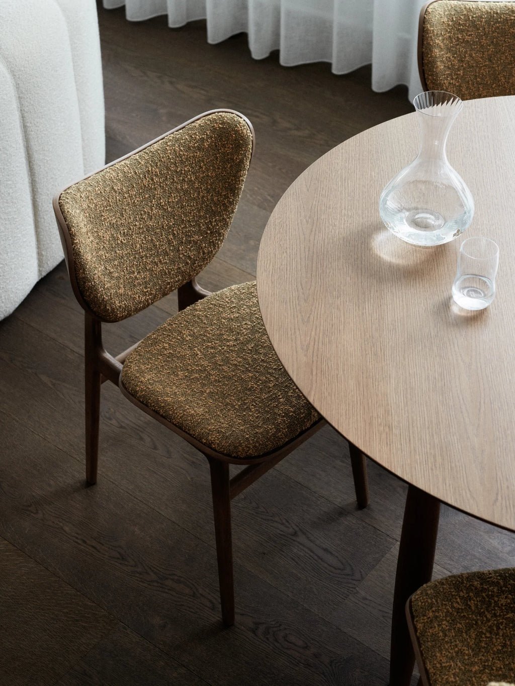 NORR11 Oku Round Dining Table, a modern oak dining table with a smooth, natural wood grain finish. This elegant and stylish table is complemented by textured, earthy-toned chairs, perfect for sophisticated dining spaces. Ideal for intimate gatherings and everyday meals, this set offers durable craftsmanship for discerning homeowners.