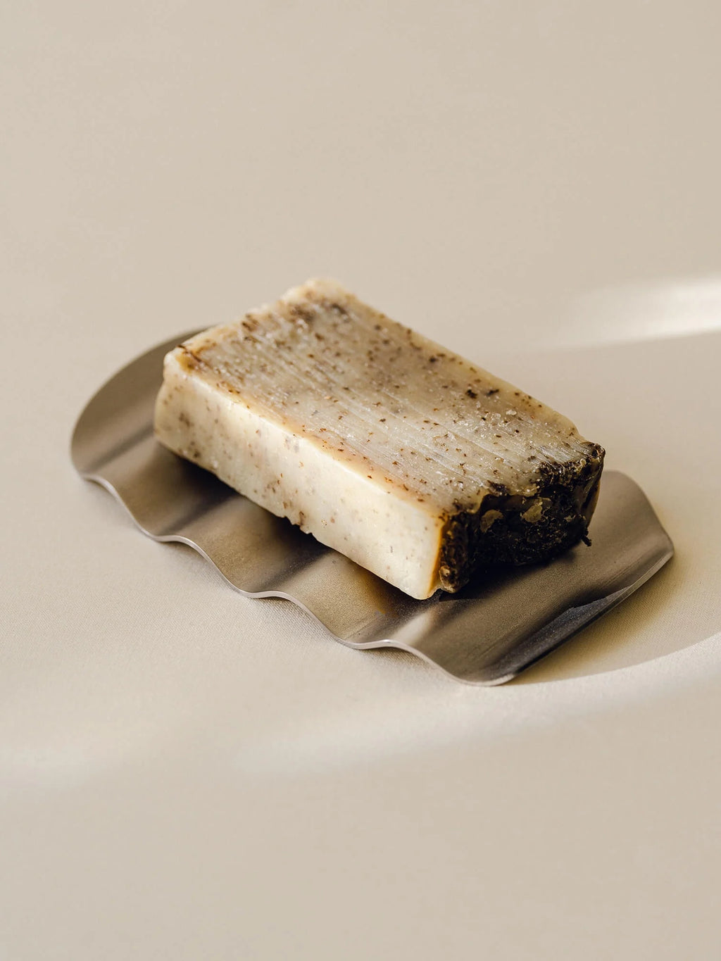 noo.ma Olbi Soap Dish, a modern minimalist soap dish crafted from brushed stainless steel, featuring a distinctive wavy, organic shape in a sleek silver finish. It elegantly holds a natural bar of soap with an off-white and speckled texture, perfect for contemporary bathroom shelf styling or adding an organic modern touch to any washroom.