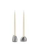 A pair of Out of Lines Candle Holders made of brushed silver or pewter metal, each with a smooth, domed base. They hold two slim, unlit white taper candles. The holders are photographed side-by-side on a white background.