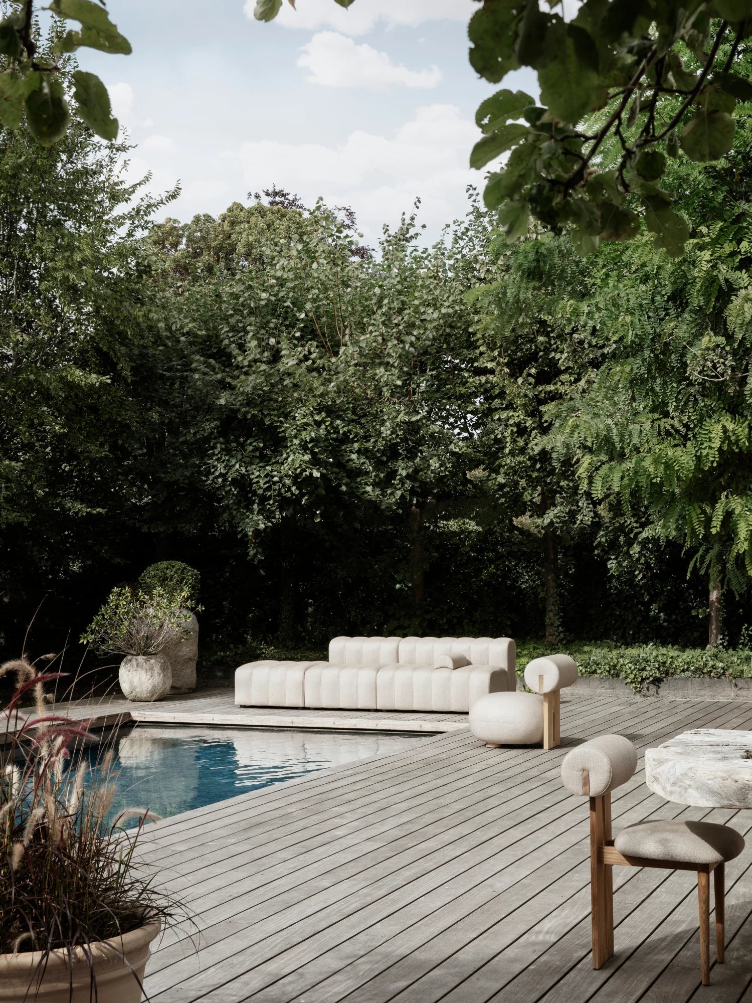 NORR11 Outdoor Hippo Lounge Chair, a sculptural organic modern outdoor lounge chair featuring plush light beige textured bouclé upholstery with a unique cylindrical backrest, set on a light natural wood frame with subtle brass accents. This contemporary outdoor seating piece is perfect for a minimalist pool deck, patio, or sophisticated garden lounge area.
