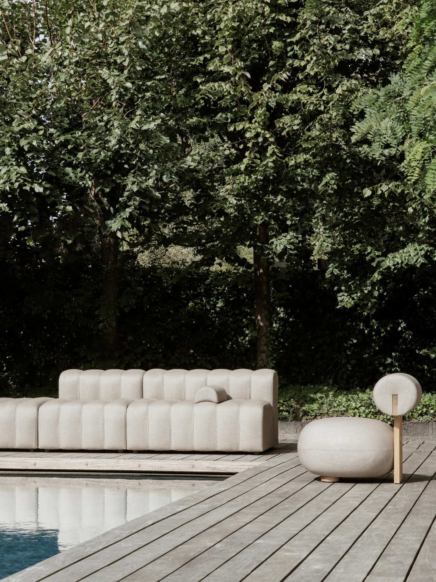 NORR11 Outdoor Hippo Lounge Chair, a striking organic modern design featuring a plump, rounded beige upholstered seat in durable textured outdoor fabric, complemented by a circular backrest on a vertical natural light wood support. This minimalist and contemporary patio chair offers stylish comfort with its distinctive silhouette. Ideal for poolside relaxation, modern gardens, or Scandinavian-inspired outdoor living areas.