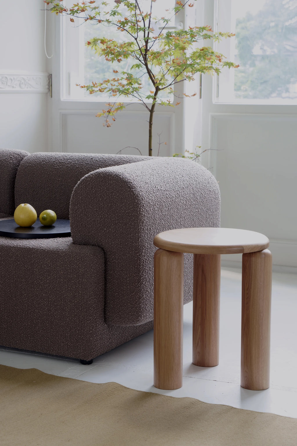TAMO Neo Side Table, a round, genuine light oak wood modern side table with three cylindrical legs, displayed next to a textured gray sofa. Enhances a contemporary living space.