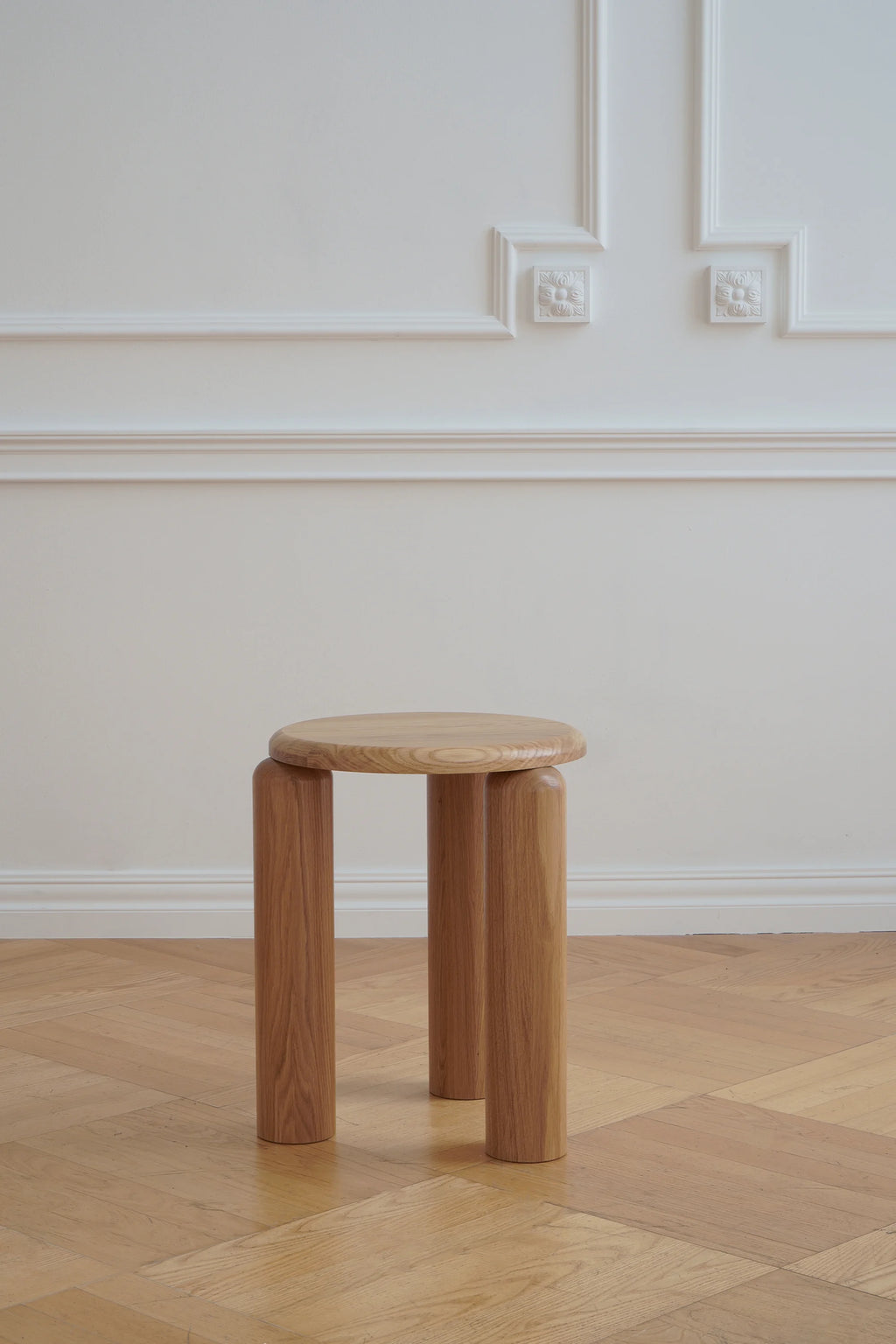 TAMO Neo Side Table in a lifestyle shot. This round, solid light oak wood mid-century modern side table features three thick cylindrical legs, displayed on a parquet floor. A classic addition to any living space.