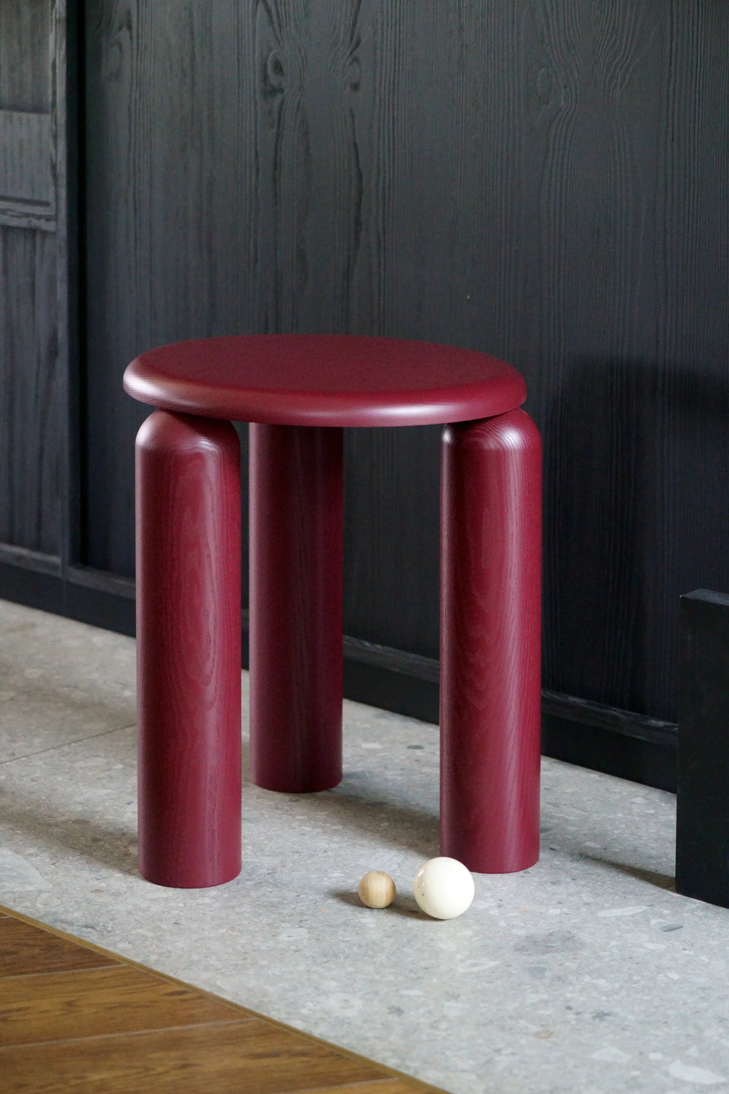TAMO Neo Side Table in a rich, dark magenta-red wood, featuring a round top and three thick, rounded legs, set against a dark textured wall. This premium, modern design offers versatile accent furniture.